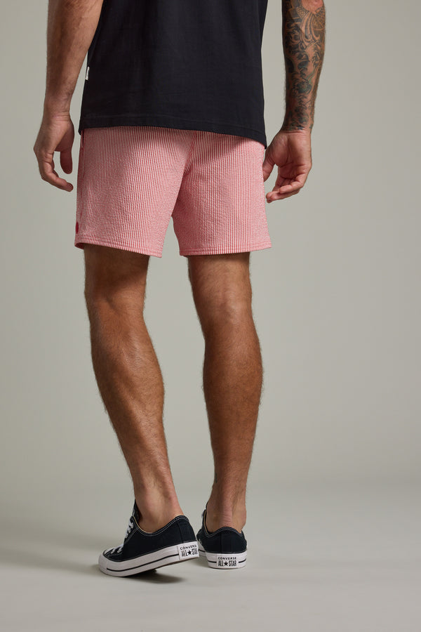 A man faces away in a black T-shirt, Barkers Dehaven Stripe Volley seersucker shorts, and black low-top sneakers, revealing a tattoo on his right forearm. The backdrop is plain and neutral.