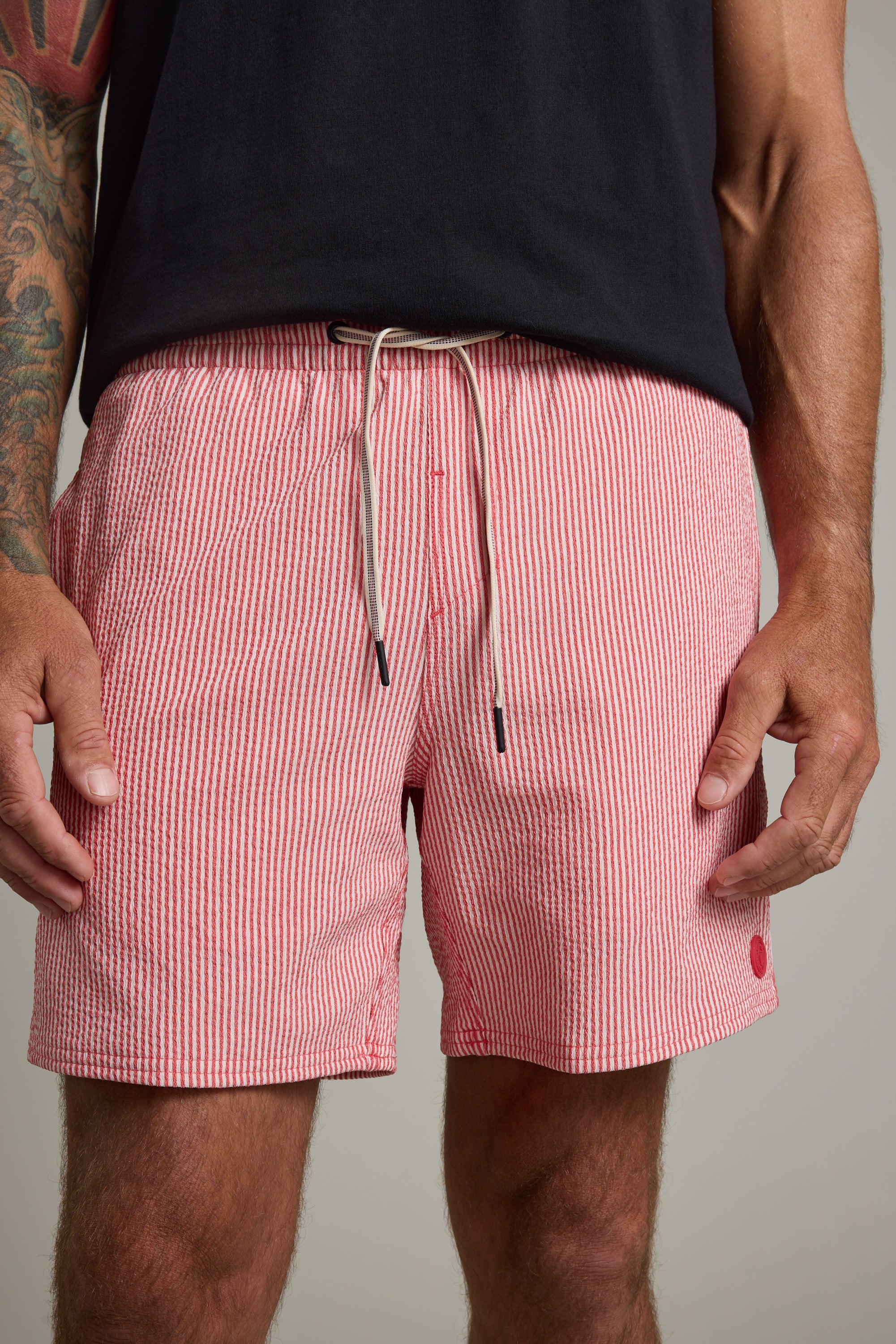 A man wears Barkers Dehaven Stripe Volley red and white seersucker shorts with a drawstring, paired with a black T-shirt. His hands rest at his sides and part of a tattoo is visible on his left arm.