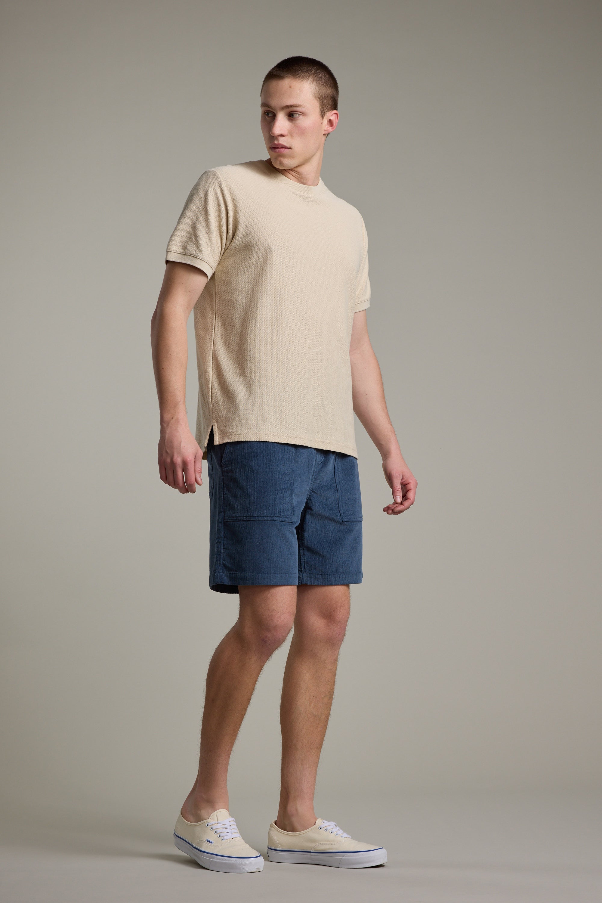 A young man stands against a neutral background, wearing a beige short-sleeve shirt and Barkers Montauk Cord Weekend Short in blue cotton corduroy with an elastic waistband. He looks left, relaxed, in beige canvas sneakers.