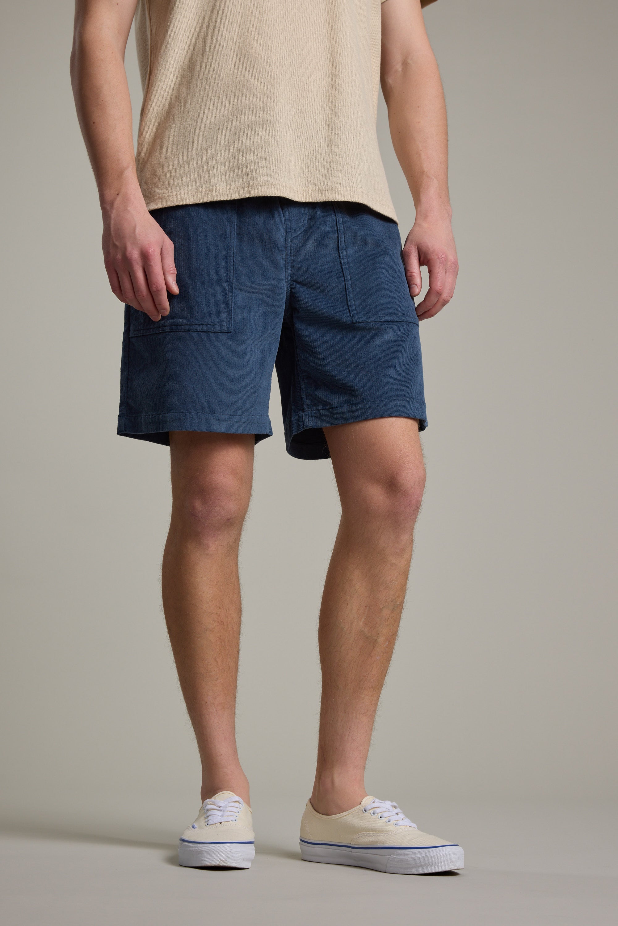 A person stands against a plain background wearing a beige short-sleeve shirt, Barkers Montauk Cord Weekend Short in blue with an elastic waistband and front pockets, and white sneakers. The image is cropped from shoulders to feet.