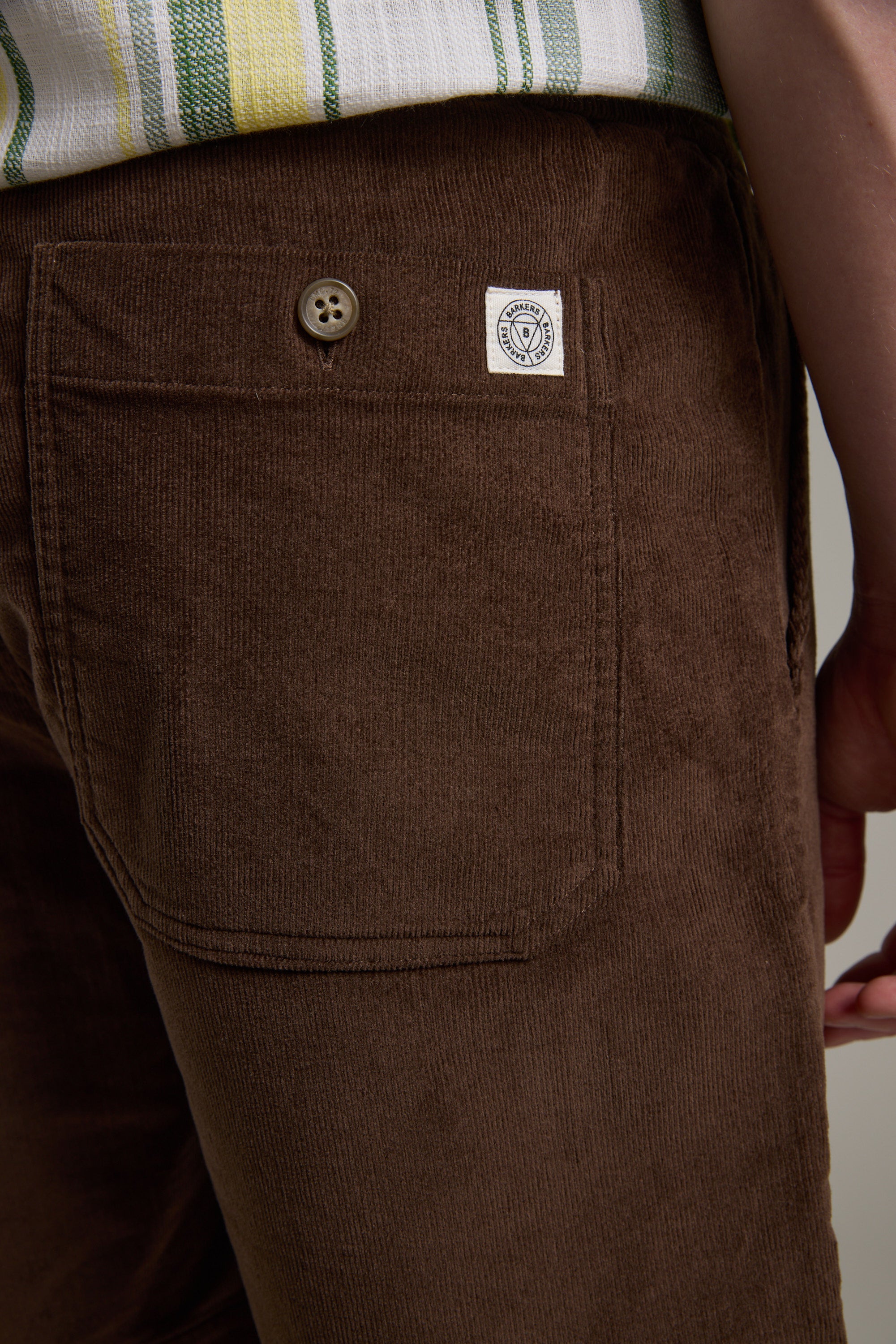 Close-up of a person wearing Barkers Montauk Cord Weekend Short in brown corduroy with a back pocket, button, and white patch label. A striped shirt in green, yellow, and white is partially visible at the waist.