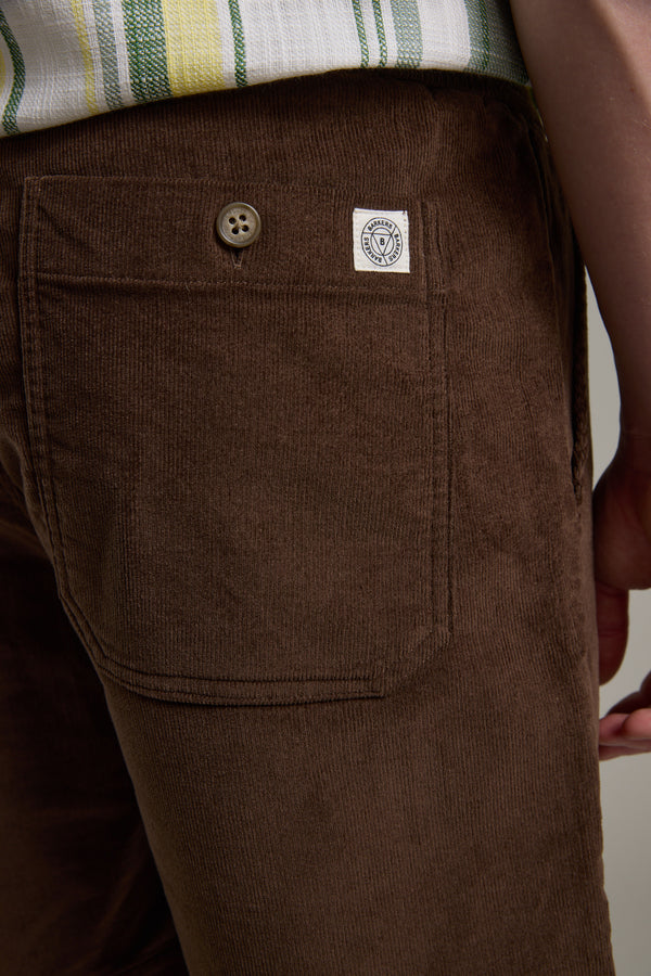 Close-up of a person wearing Barkers Montauk Cord Weekend Short in brown corduroy with a back pocket, button, and white patch label. A striped shirt in green, yellow, and white is partially visible at the waist.