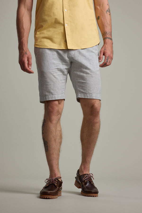 Wearing Barkers' Palma Walkshort in light gray, a person with visible arm and leg tattoos stands relaxed, paired with a yellow short-sleeve shirt and brown boat shoes.
