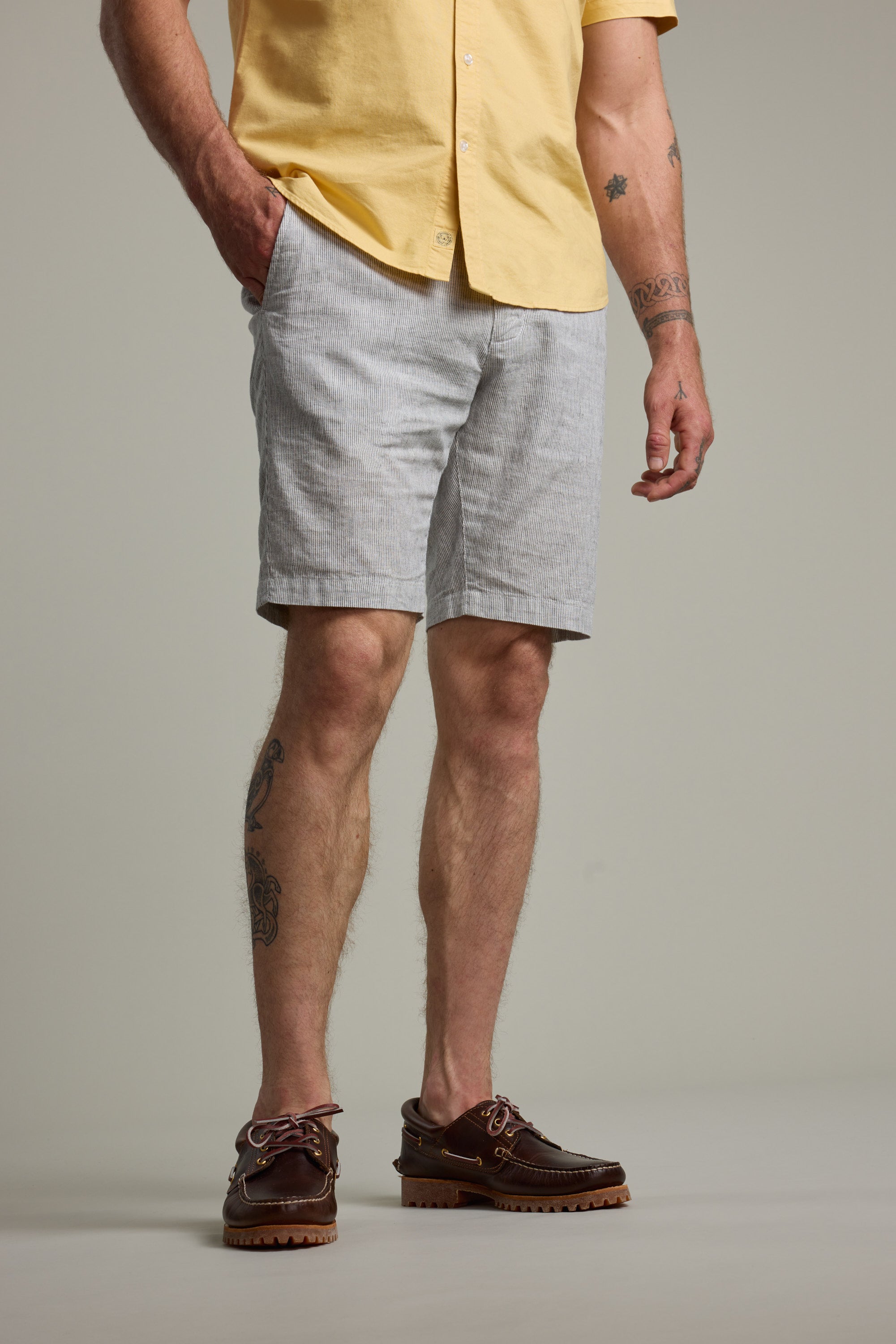 A man with visible forearm and leg tattoos stands with one hand in his pocket, wearing a yellow short-sleeve shirt, Barkers Palma Walkshorts in light gray linen-cotton, and brown boat shoes.