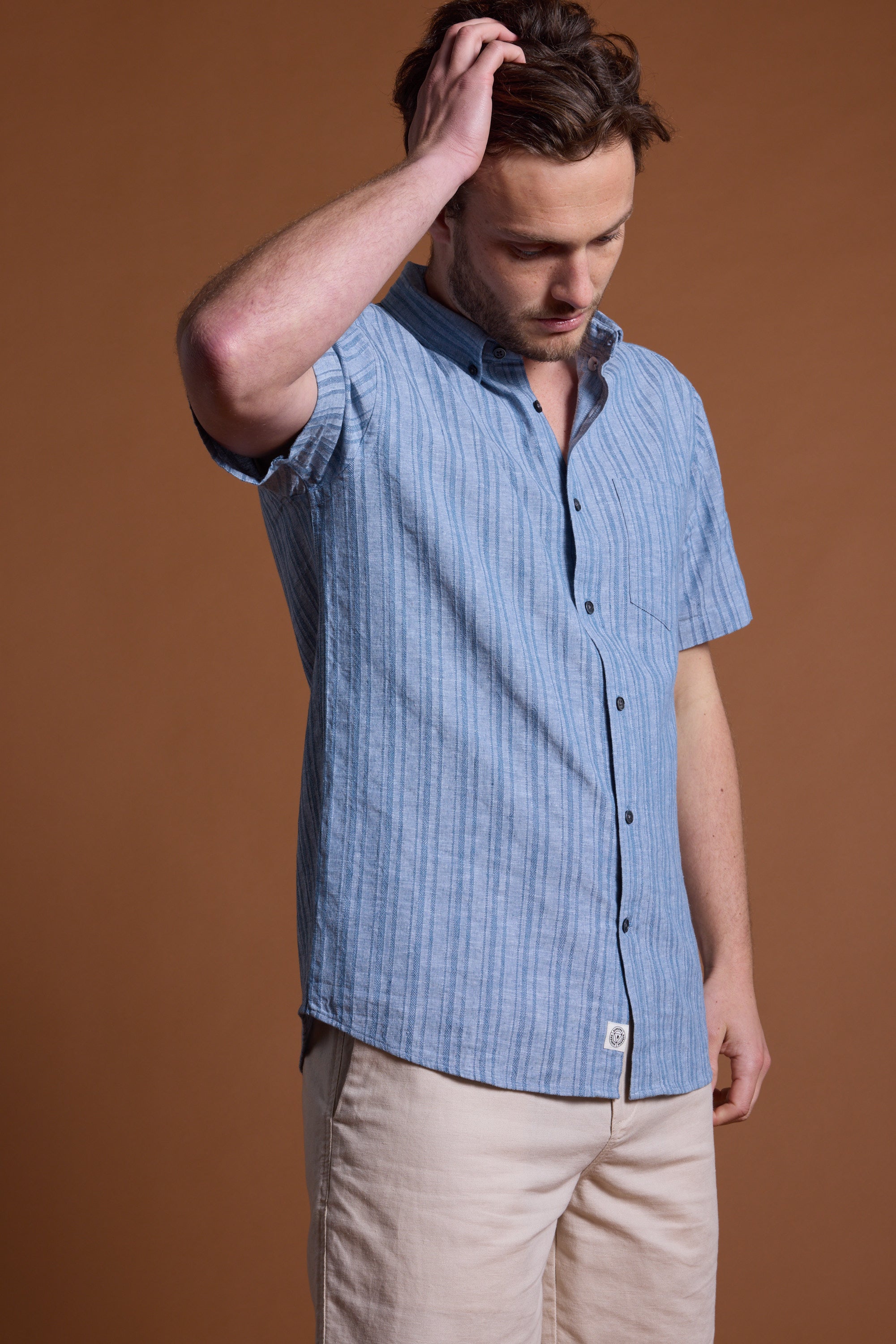 A man in the Barkers Garvey Stripe Shirt and beige pants stands against a brown background, looking down while touching his hair—the perfect summer essential.
