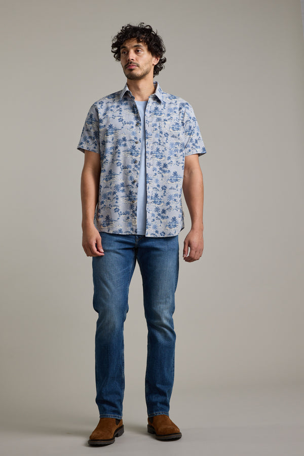 A man stands against a plain background wearing the Barkers Radford Print Shirt, a light blue Hawaiian shirt made from responsibly sourced cotton, layered over a white T-shirt with blue jeans and brown shoes. He faces slightly left with a neutral expression.