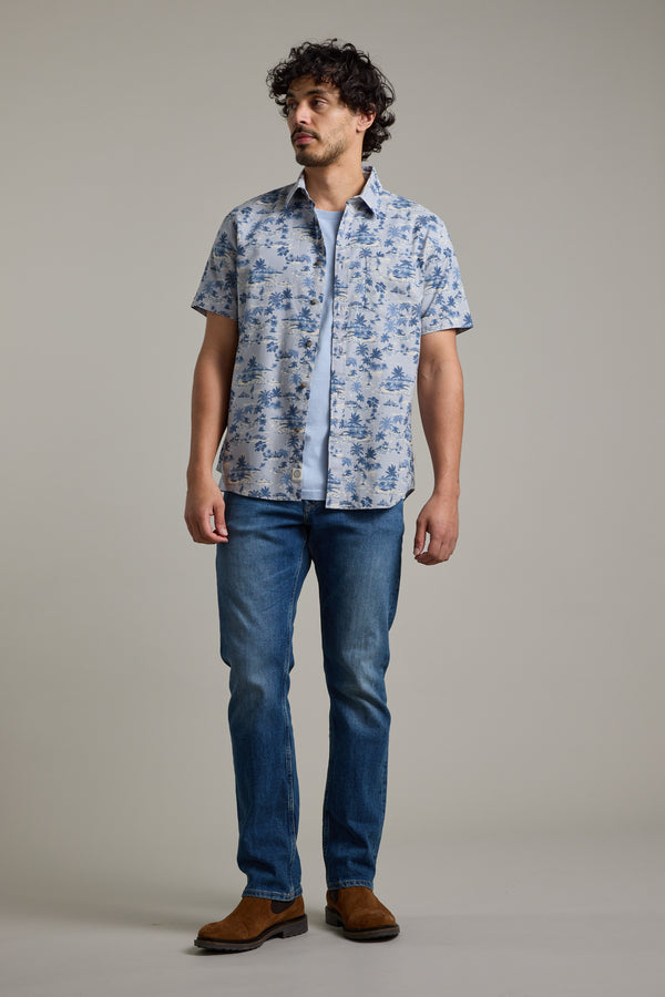A man with curly hair models the Barkers Radford Print Shirt—a tonal blue Hawaiian shirt made from responsibly sourced cotton—over a white t-shirt, blue jeans, and brown boots, standing against a plain light gray background.
