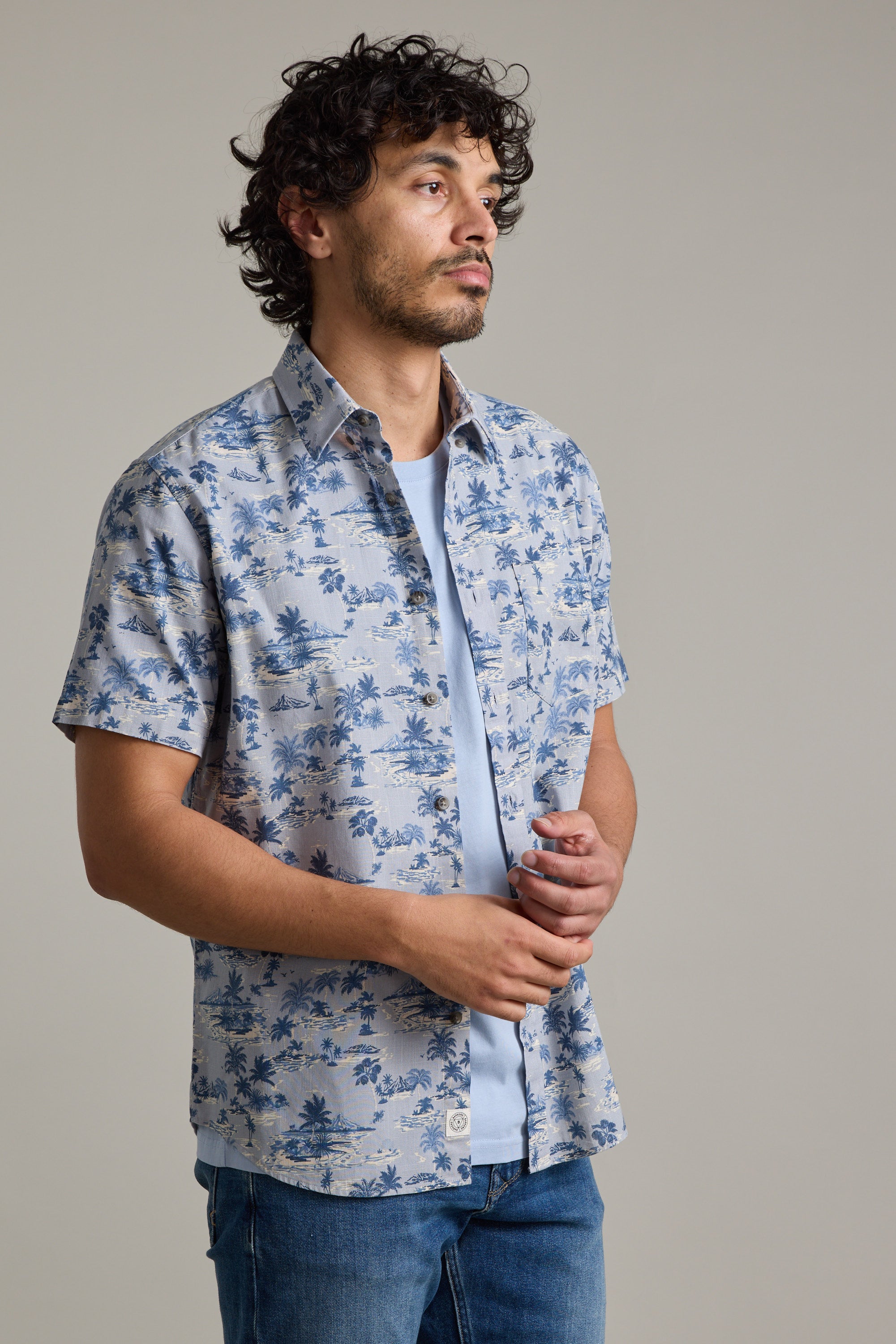 A man with curly hair and a beard wears the Barkers Radford Print Shirt, made from responsibly sourced cotton, over a T-shirt and blue jeans. He stands against a plain grey background, looking off to the side.