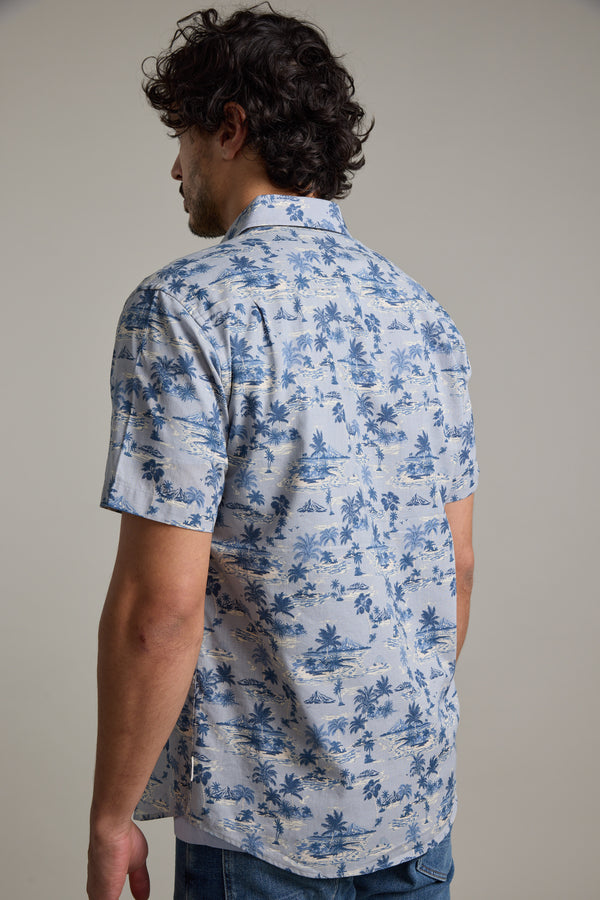 A man with curly dark hair, seen from behind, wears the Barkers Radford Print Shirt—a light blue men’s summer shirt with a tropical palm tree design—paired with blue jeans, standing against a plain light gray background.
