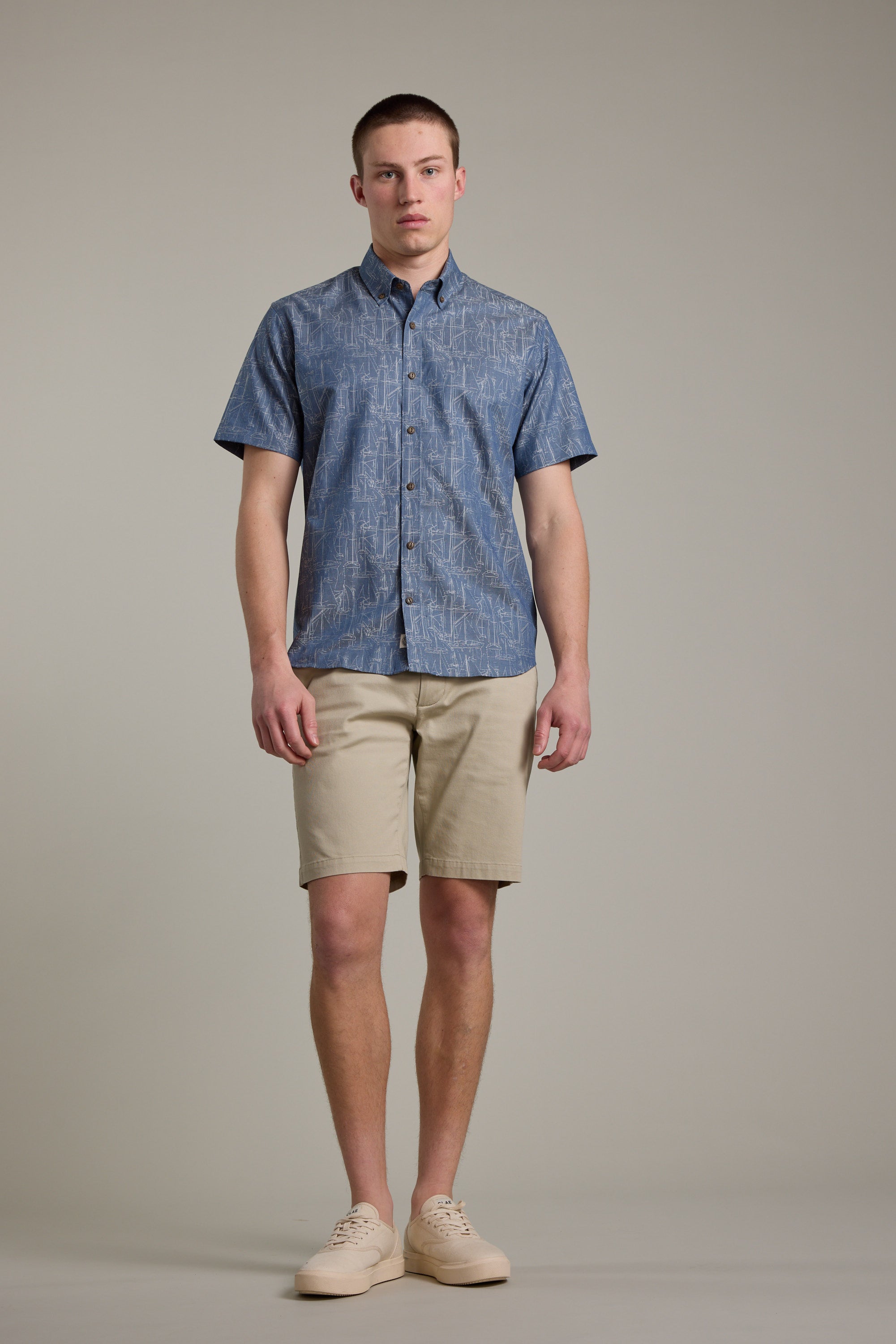 A man faces forward with a neutral expression, wearing the Barkers Yacht Jacquard Shirt against a plain background. He pairs the blue patterned summer shirt with beige shorts and light-colored sneakers.