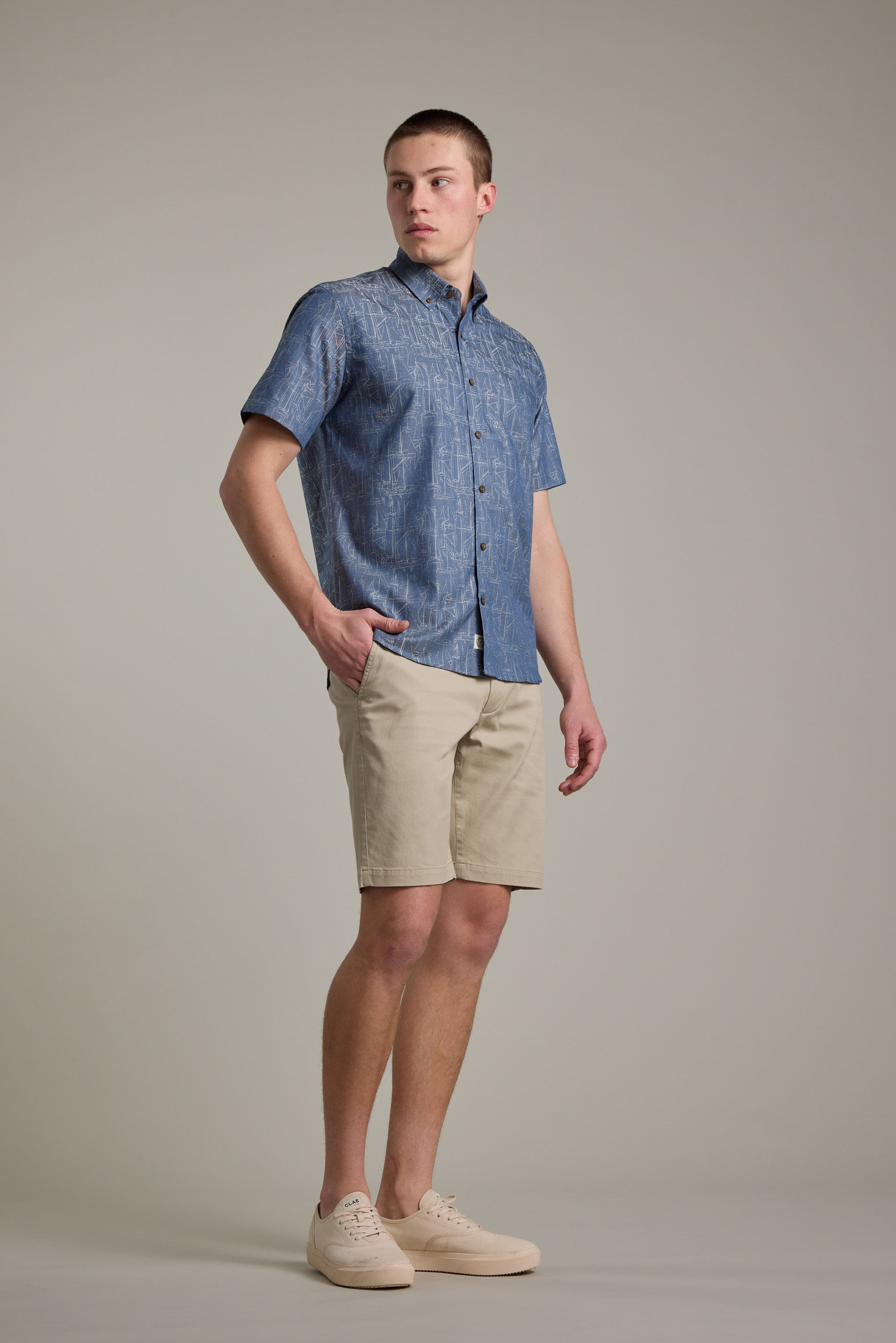 A person stands against a plain background wearing the Barkers Yacht Jacquard Shirt with beige shorts and light sneakers, one hand in their pocket and looking to the side.