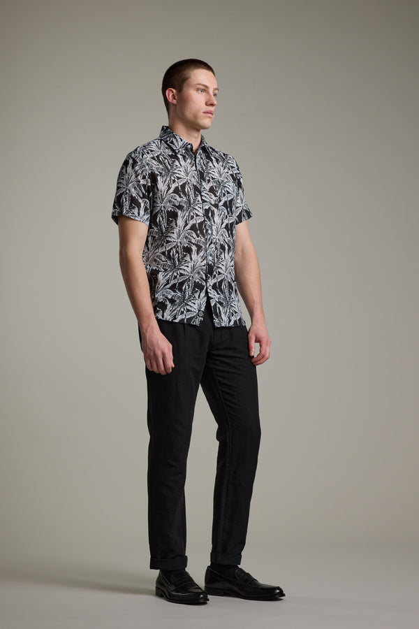 A man stands against a plain background, wearing the Barkers Madison Print Shirt—a black and white Hawaiian style in a cotton–linen blend—paired with black pants and dress shoes, looking slightly to the side.