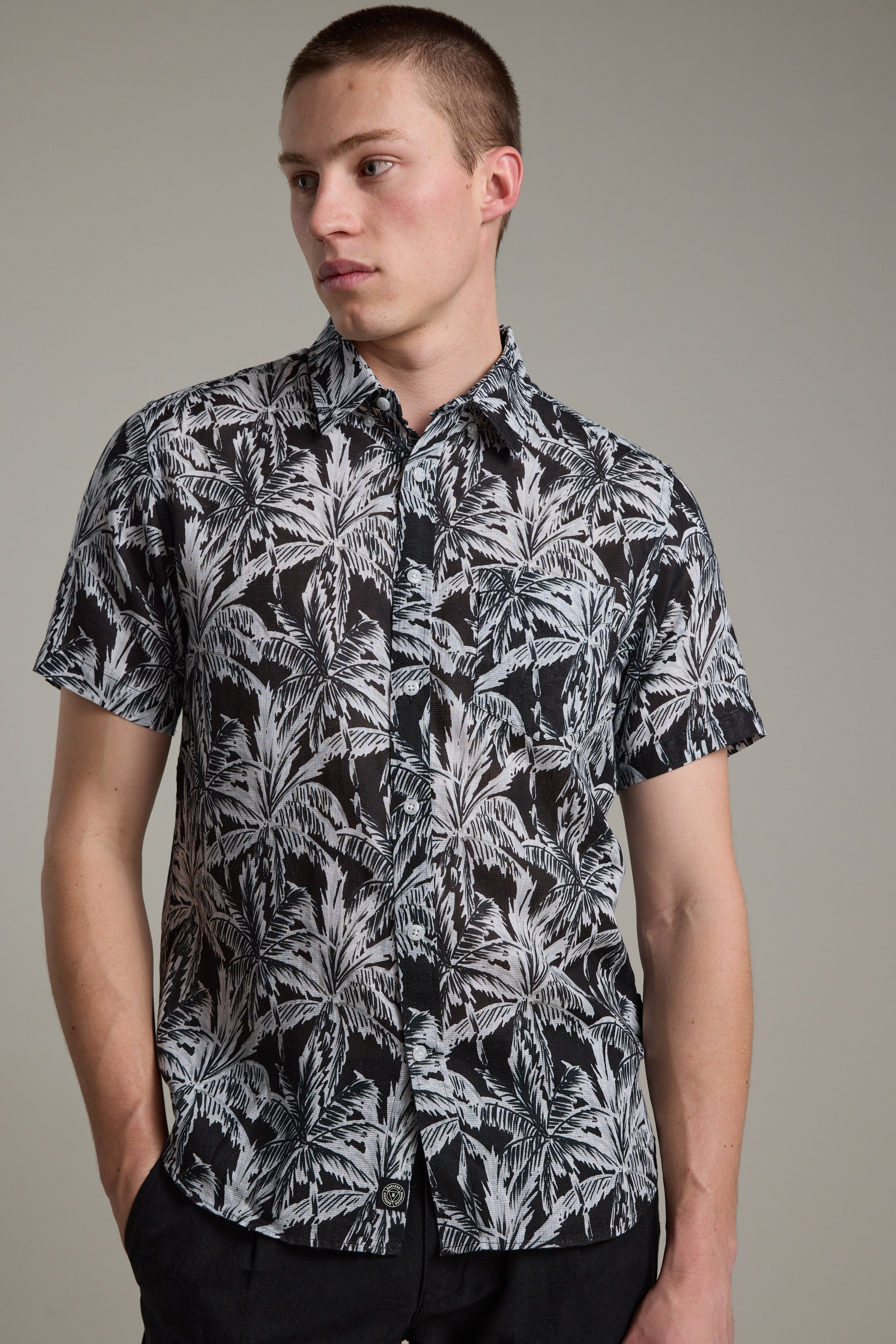 A young man with short hair models the Barkers Madison Print Shirt—a black-and-white palm leaf design in a linen-cotton blend. He stands against a plain background, one hand in his pocket and gazing to the side.