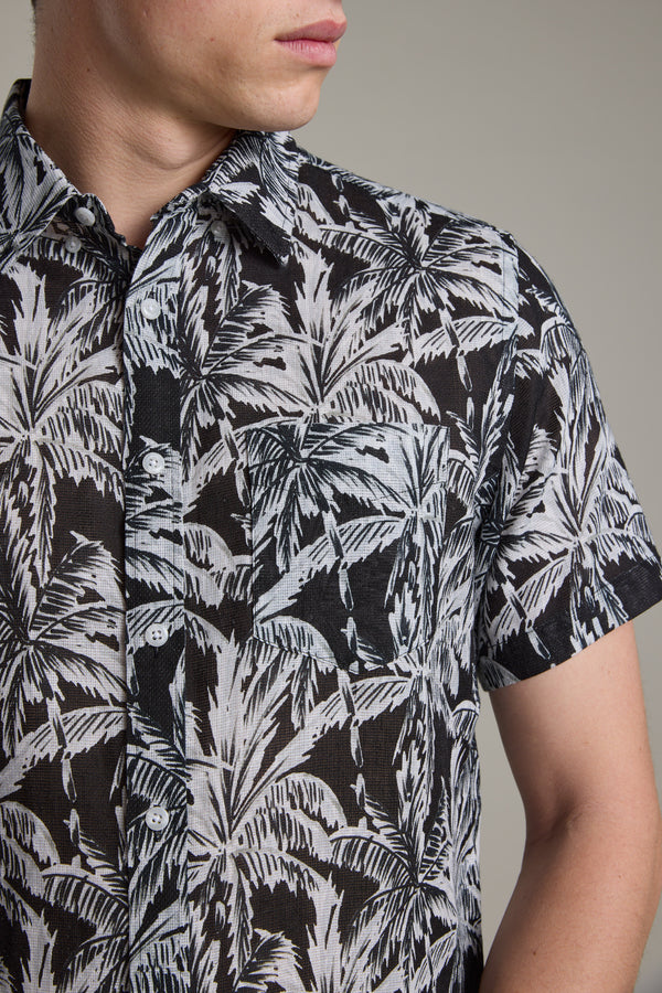 A person wears the Barkers Madison Print Shirt, a short-sleeved black Hawaiian shirt with a white palm leaf pattern, shown from shoulders to mid-torso, face partially visible and looking left.