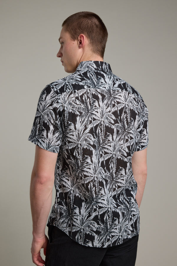 A man faces away, wearing Barkers' Madison Print Shirt—a short-sleeve black Hawaiian shirt with a white palm leaf pattern in soft cotton-linen blend—paired with dark pants against a plain light gray background.