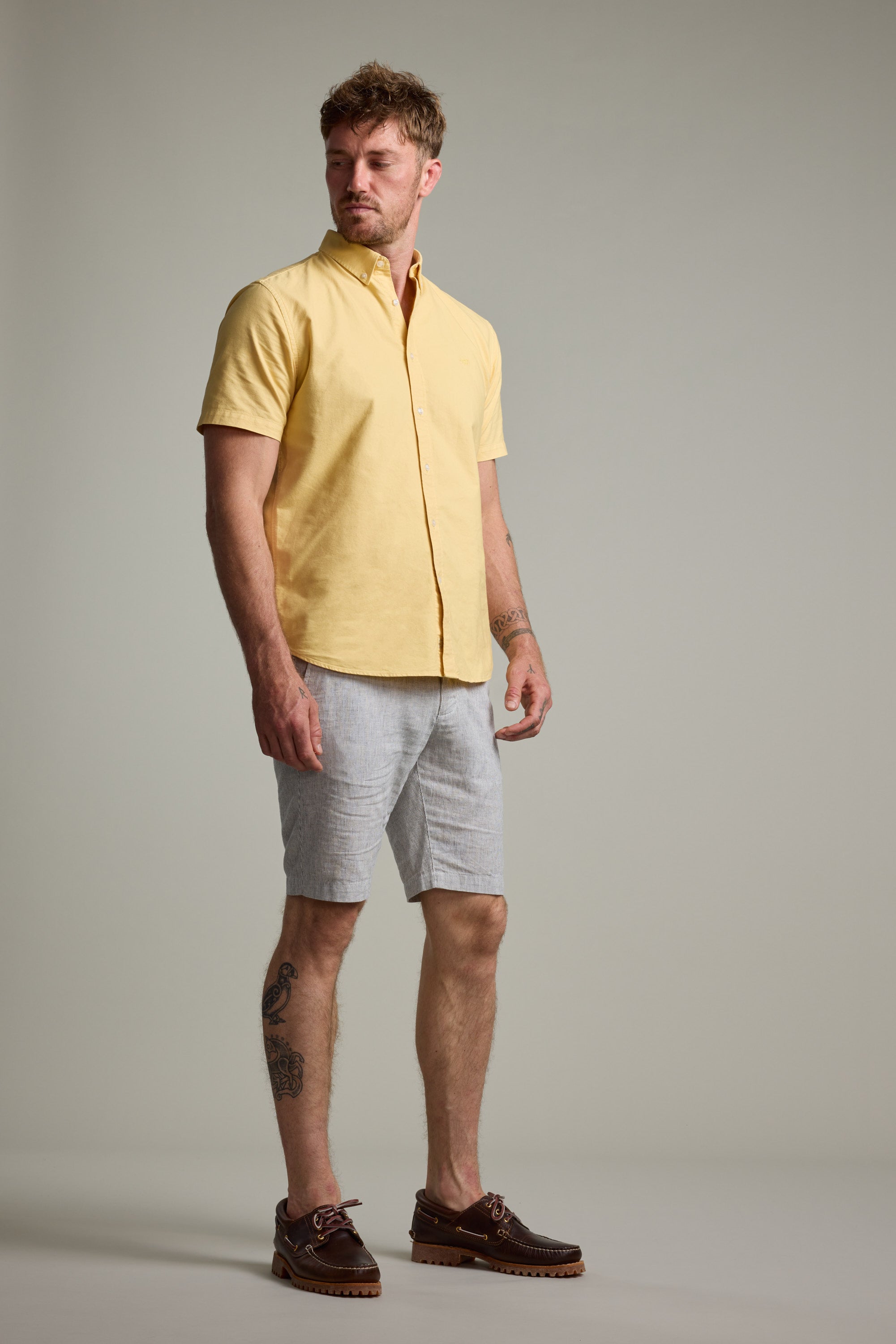 A man stands against a plain background wearing Barkers' Palma Walkshorts in light gray, a short-sleeve yellow button-up shirt, and brown boat shoes. He has short hair, leg tattoos, and looks slightly to the side.