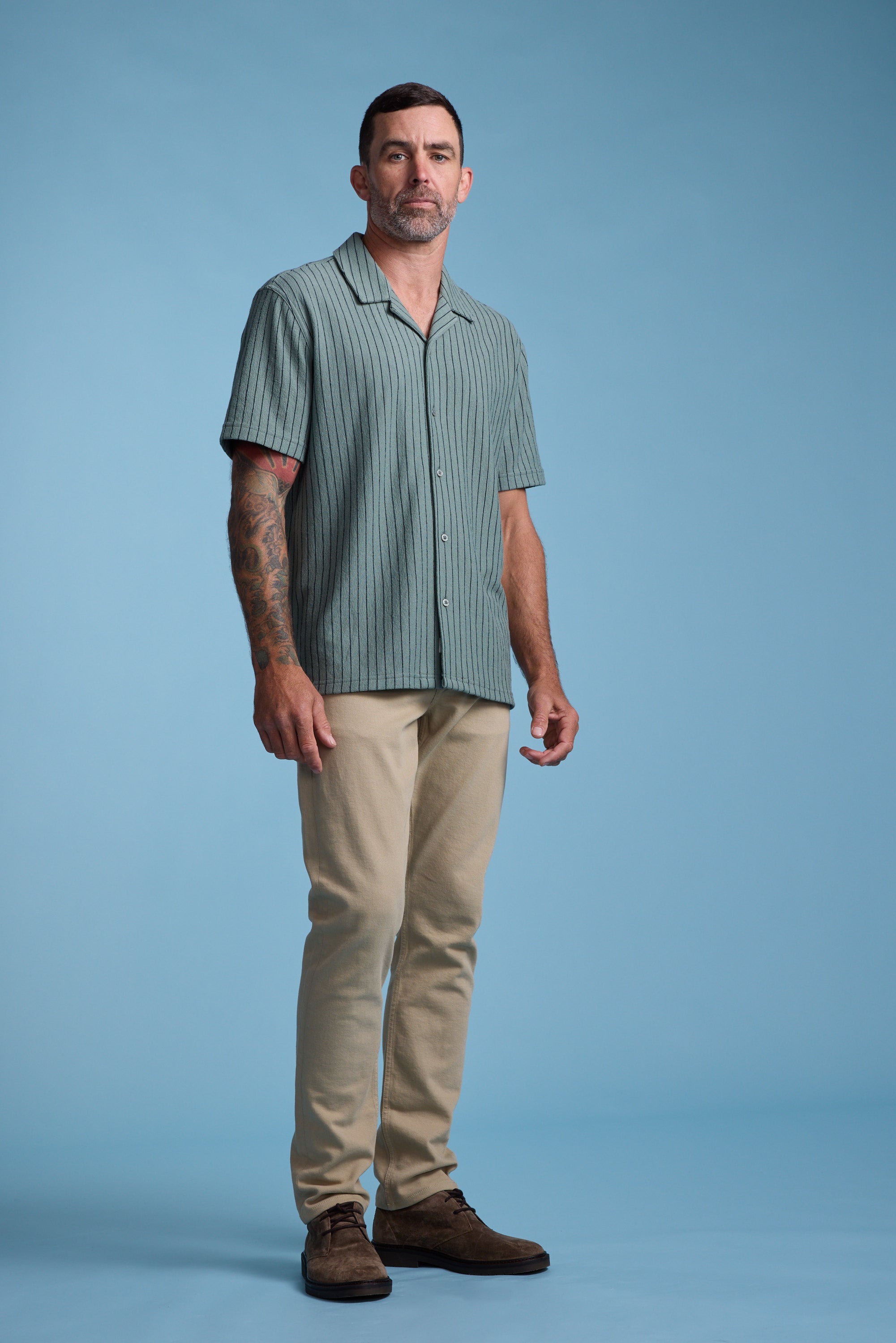 A man with short dark hair and tattoos on his right arm stands against a blue background, wearing the Barkers Diego Stripe Cuban Shirt, which has a boxy fit, paired with beige pants and brown shoes.