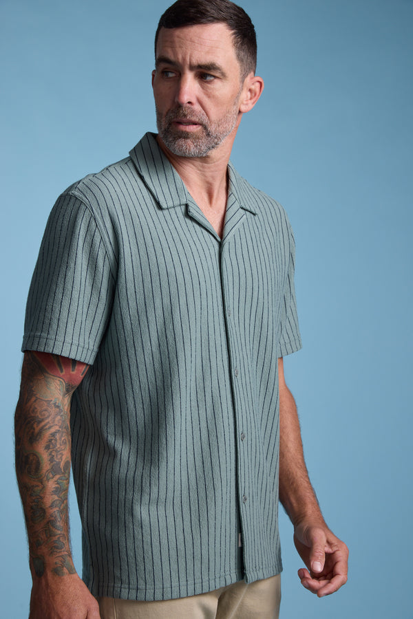 A man with short dark hair, a beard, and a tattooed arm stands against a solid blue background wearing the Barkers Diego Stripe Cuban Shirt in light green and beige pants.