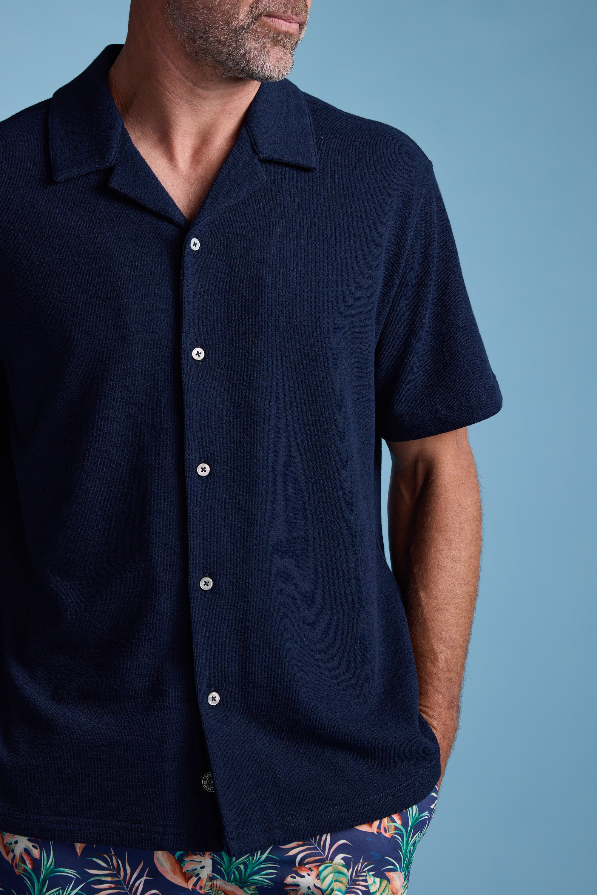 A man in the Barkers Diego Cuban Shirt and colorful tropical shorts stands against a light blue background with one hand in his pocket; his face is partially out of frame.