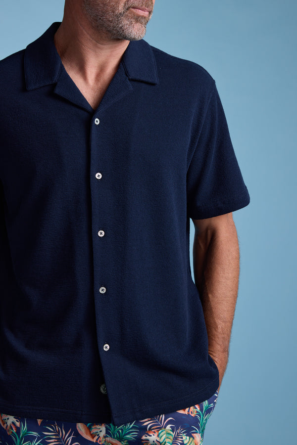 A man in the Barkers Diego Cuban Shirt and colorful tropical shorts stands against a light blue background with one hand in his pocket; his face is partially out of frame.
