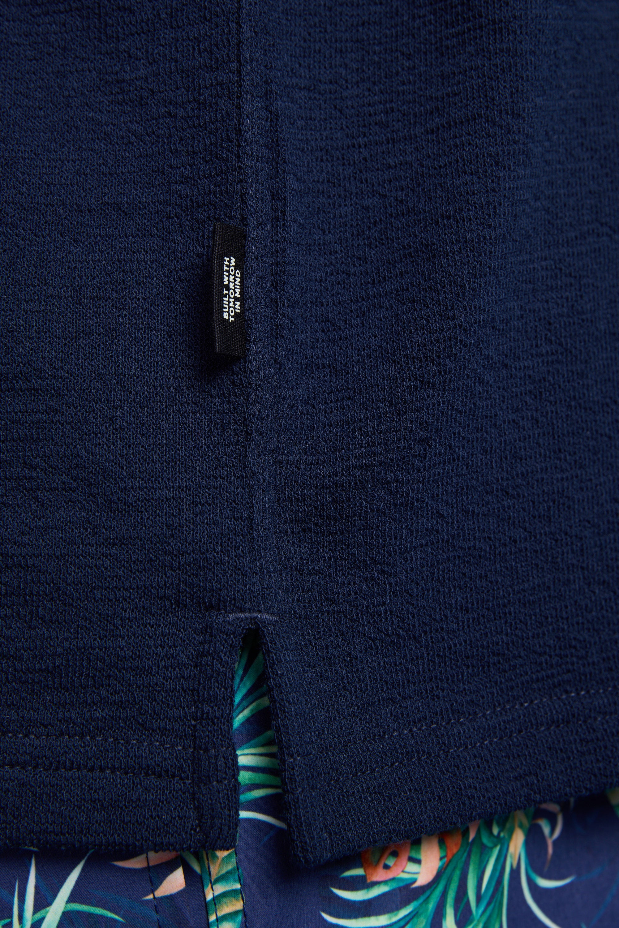 A close-up of the Barkers Diego Cuban Shirt in navy textured terry toweling with a boxy fit and small black tag near the hem, paired with blue shorts featuring a green and orange tropical leaf pattern.