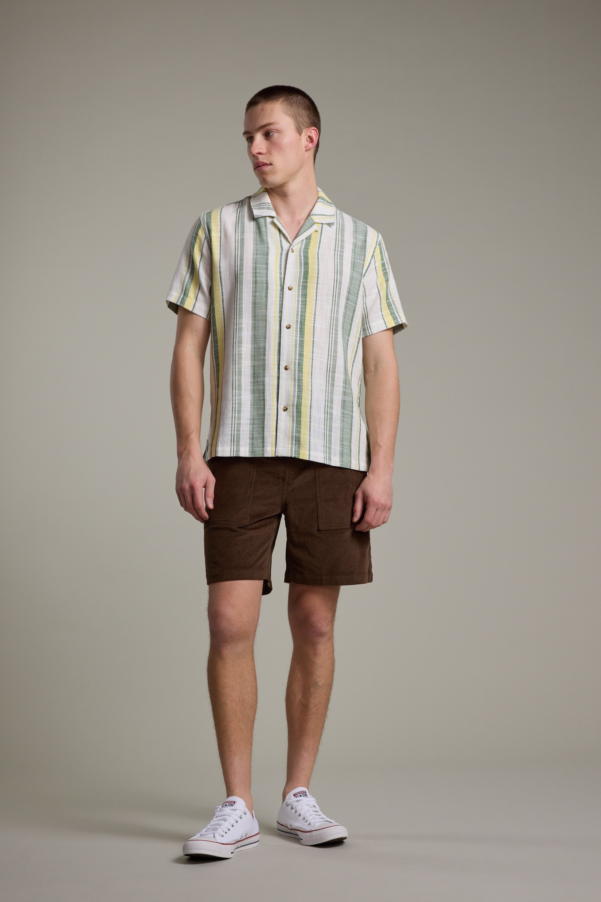 A man stands against a plain background in a short-sleeve, striped button-up shirt with green, yellow, and white tones, Barkers Montauk Cord Weekend Shorts with an elastic waistband, and white sneakers. He looks to the side, relaxed.