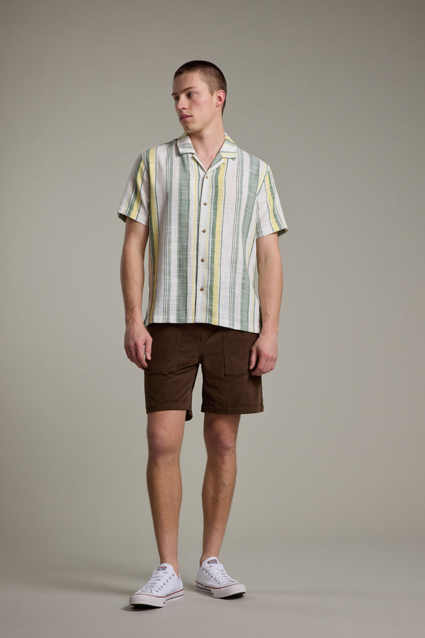 A man stands against a plain background in a short-sleeve, striped button-up shirt with green, yellow, and white tones, Barkers Montauk Cord Weekend Shorts with an elastic waistband, and white sneakers. He looks to the side, relaxed.