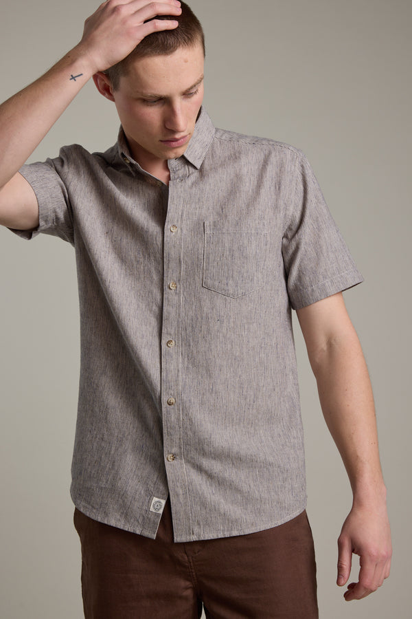 Wearing the Barkers Woodley Texture Shirt, a short-sleeve gray cotton-linen blend, and brown pants, a person stands against a plain background, looking down while touching their head with one hand.