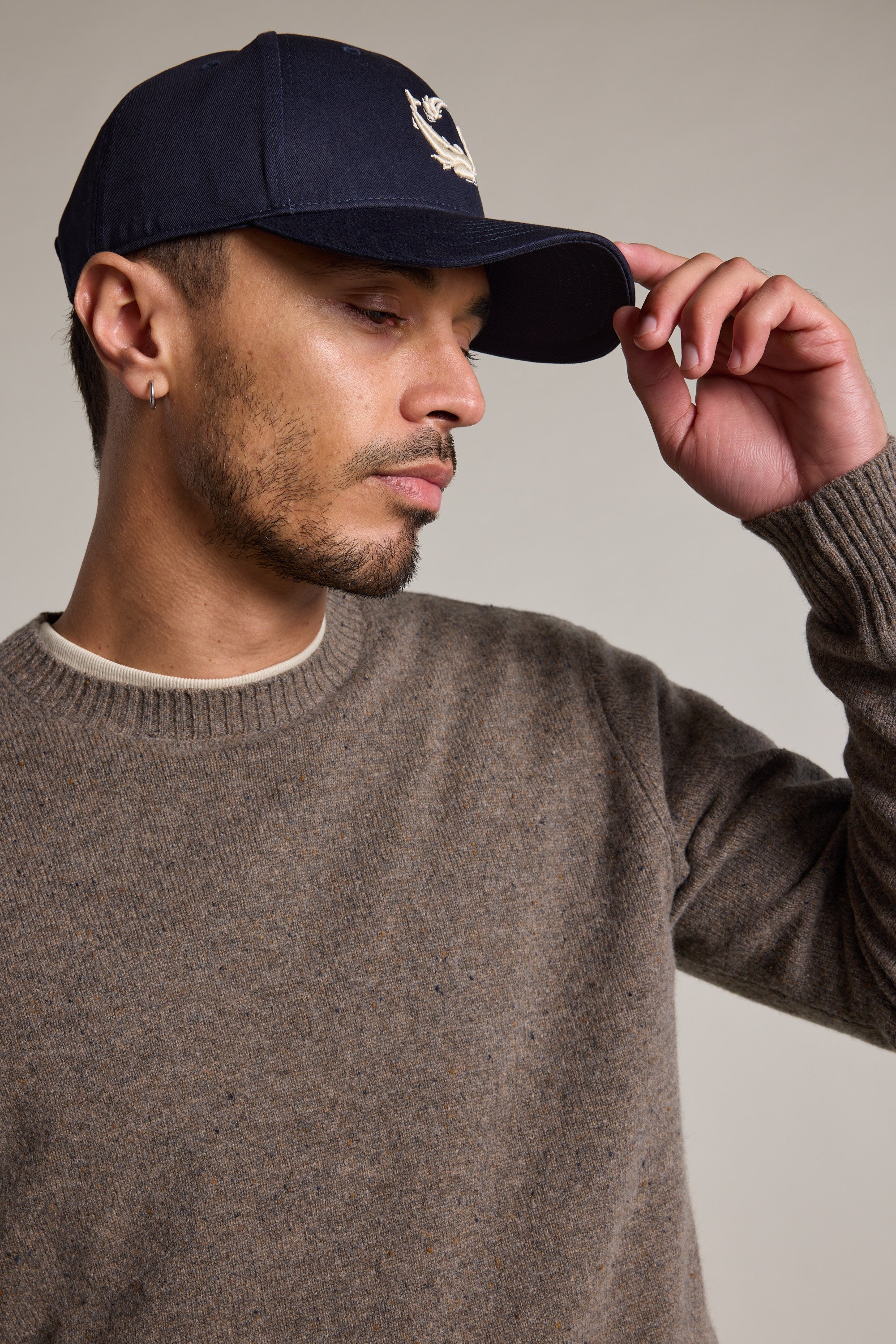 A man in a brown knit sweater wears the Legasea Logo Cap by Legasea, featuring an embroidered fish design, as he tilts the 5-panel cap with one hand and looks down to the side against a plain background.
