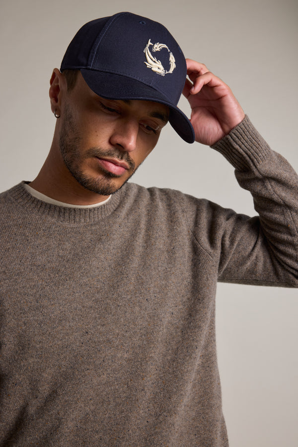 A man in a brown sweater and navy Legasea Logo Cap with an embroidered fish design lightly touches the brim of his Legasea cap, looking down with a neutral expression.