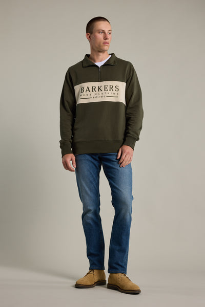 A man with short hair stands against a plain background wearing the Barkers Heritage 1/4 Zip sweater in green, paired with blue jeans and tan suede shoes, looking slightly to the side.
