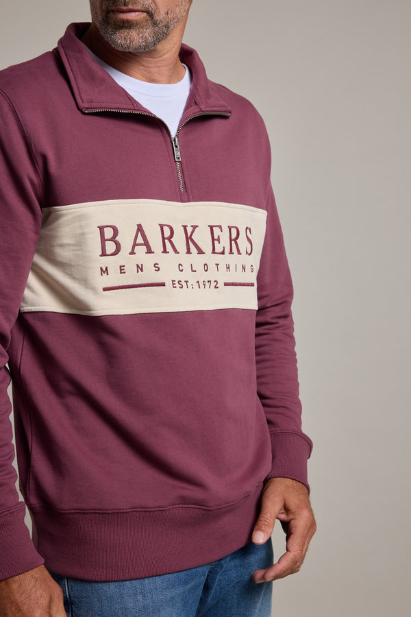 A man wears the Barkers Heritage 1/4 Zip sweater by Barkers, featuring a maroon body with a beige panel printed with "BARKERS MENS CLOTHING EST. 1972", paired with blue jeans, standing against a plain gray background.