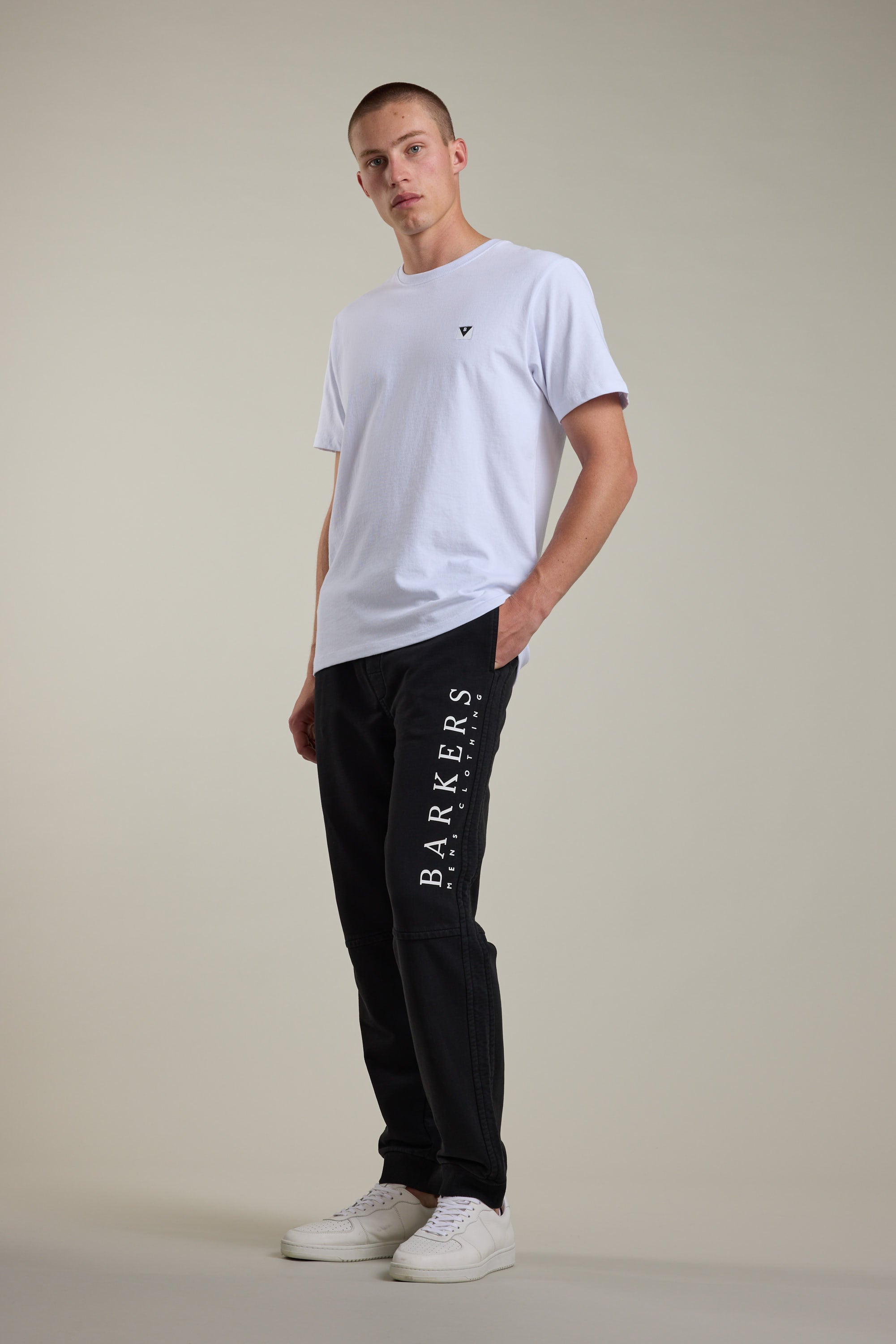 A person poses against a plain background in a white t-shirt, Barkers Organic Classic Print Track Pant with 'BARKERS' along the leg, and white sneakers—one hand in their pocket, evoking classic ‘90s trackies style.