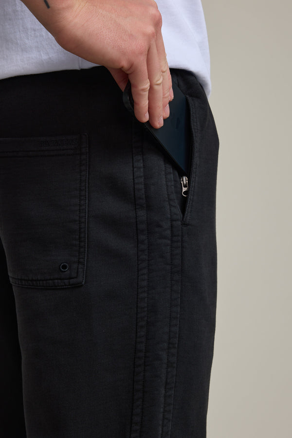 Someone places a black smartphone into the zippered pocket of their Barkers Organic Classic Print Track Pant. The person's upper body and part of a white shirt are also visible.