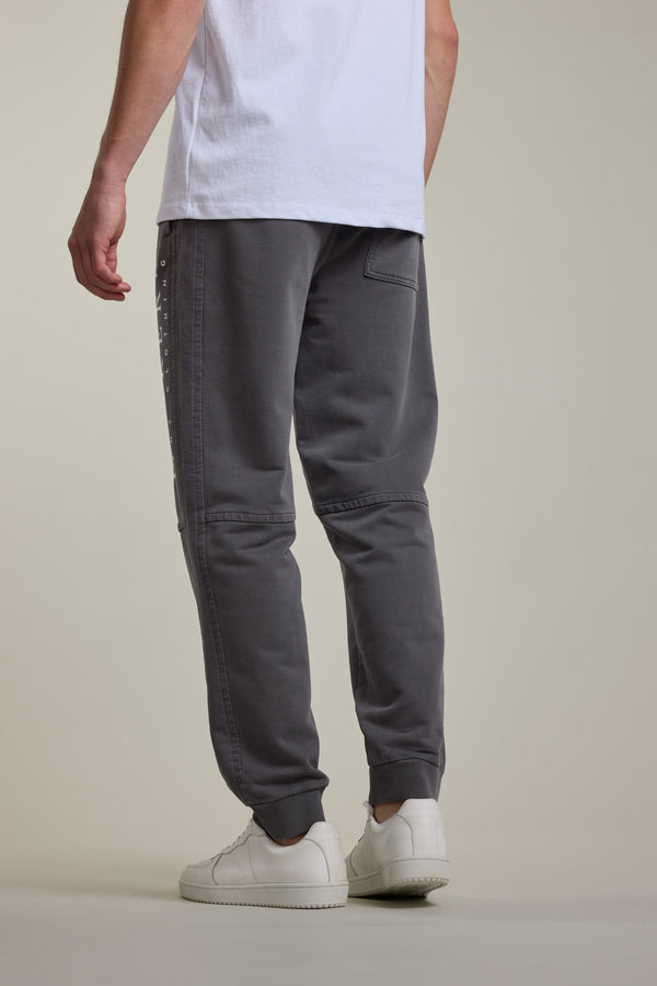 A person in a white T-shirt, Barkers Organic Classic Print Track Pant, and white sneakers stands facing away from the camera against a plain light background.