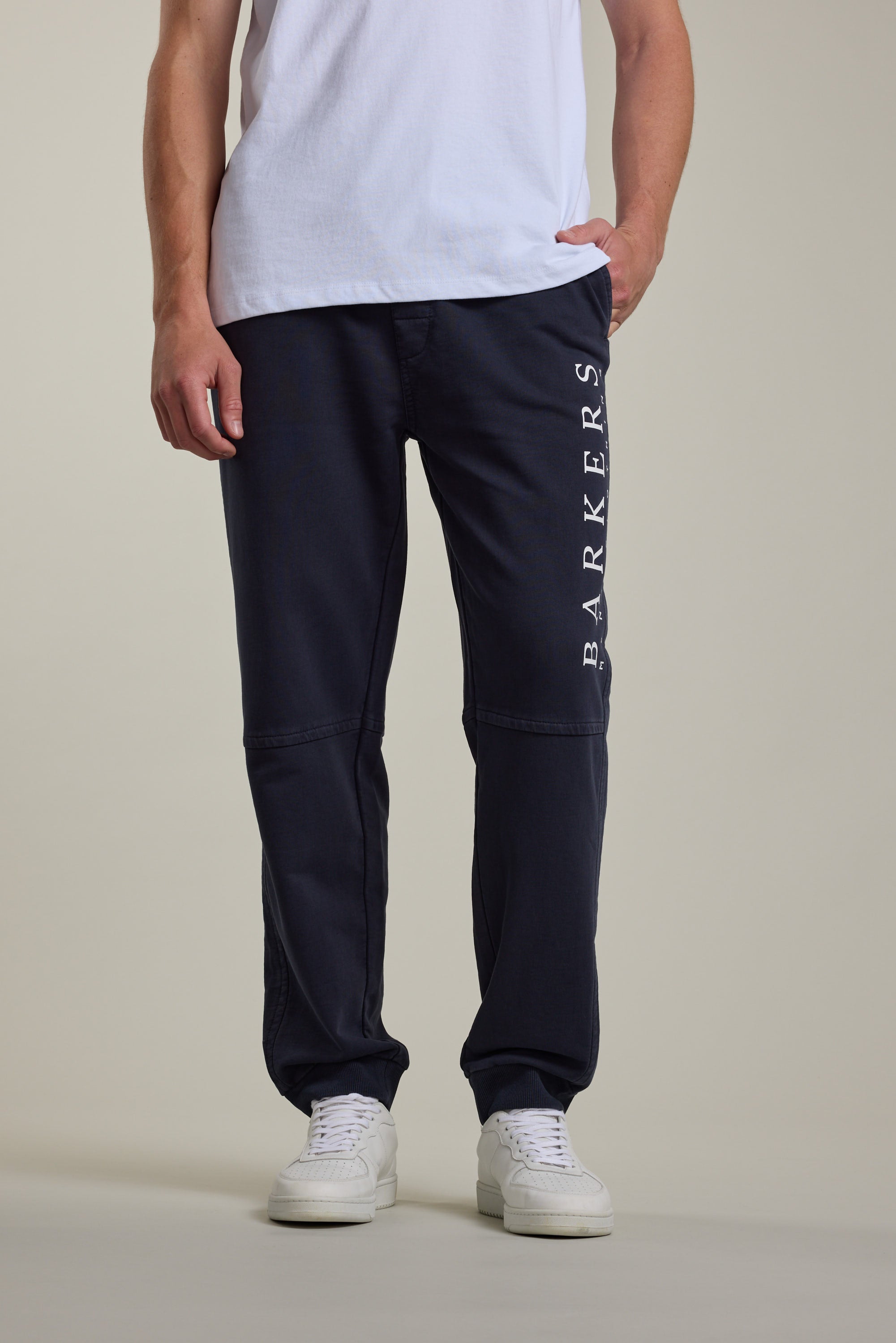 A person in a white T-shirt, Barkers Organic Classic Print Track Pant with BARKERS printed vertically on one leg, and white sneakers stands against a plain background.