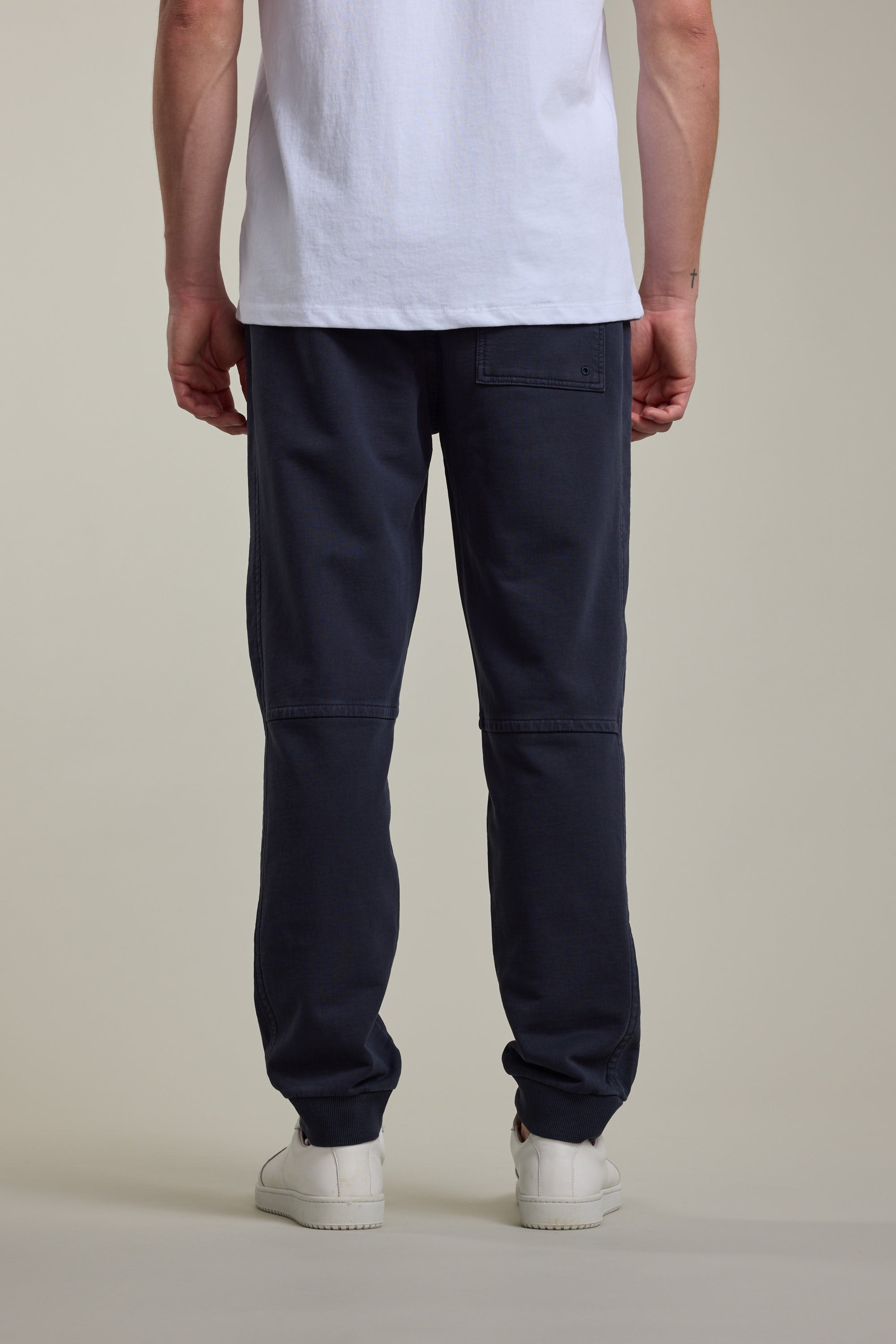 A person faces away, wearing a white T-shirt, Barkers Organic Classic Print Track Pant in dark blue with a back pocket, and white sneakers, against a plain light background.