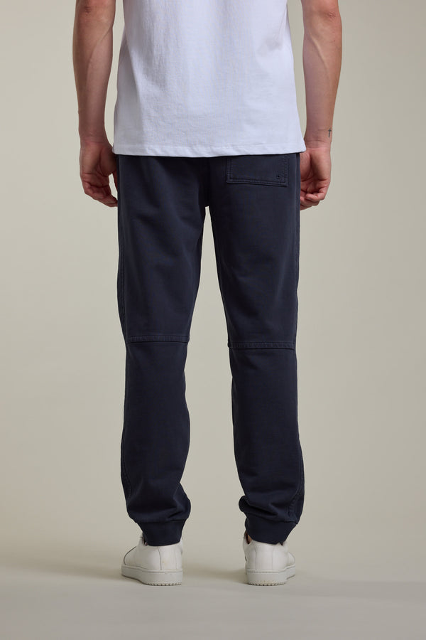 A person faces away, wearing a white T-shirt, Barkers Organic Classic Print Track Pant in dark blue with a back pocket, and white sneakers, against a plain light background.