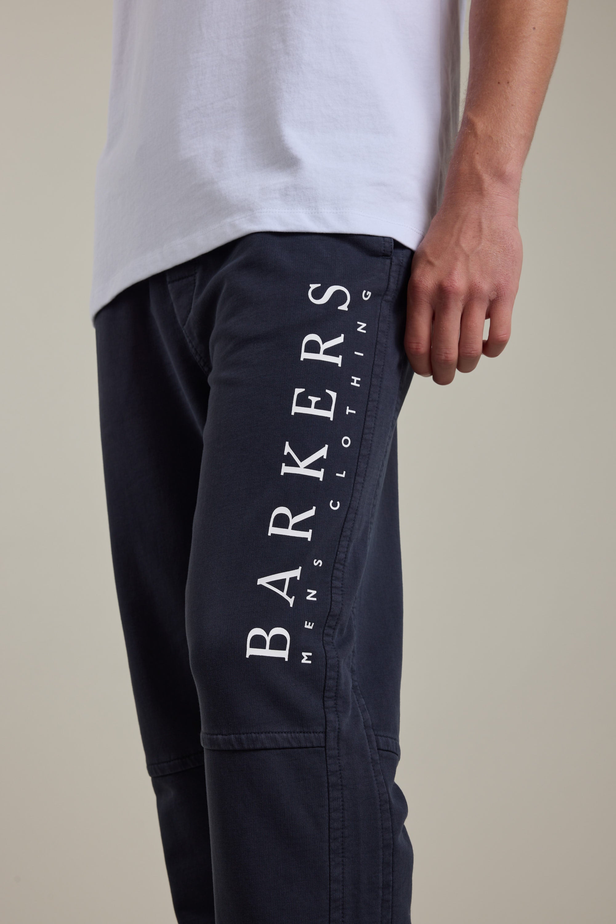 A person wearing a white t-shirt and dark blue Barkers Organic Classic Print Track Pant with "BARKERS" mens clothing printed vertically on the side of the left leg.