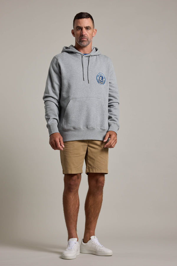A man stands facing forward against a plain light background, wearing a light gray Swordfish Mojo Logo Hoody with a front pocket and chest logo, tan shorts, and white sneakers.
