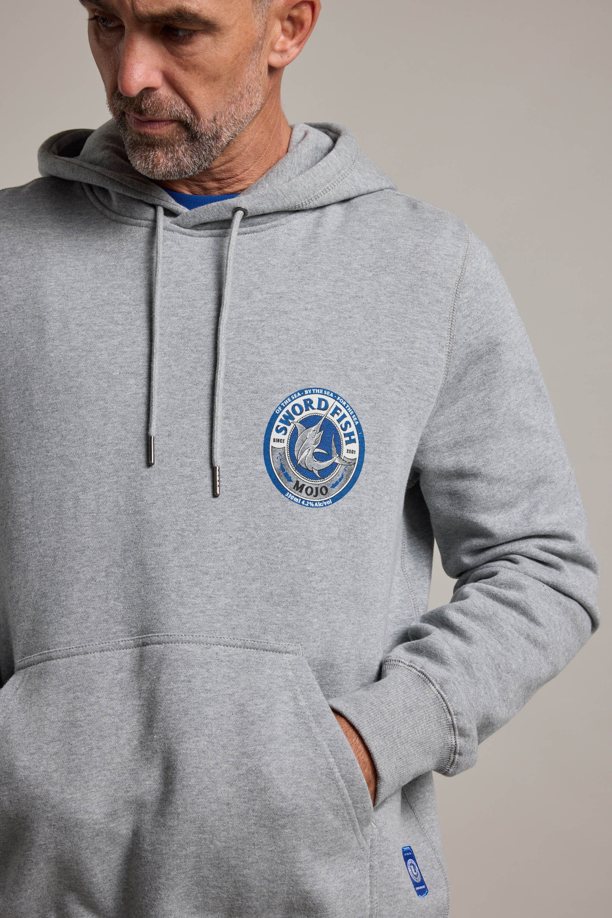 A man wears the Swordfish Mojo Logo Hoody in light gray, featuring the brand’s logo on the chest, with one hand in its front pocket. Ideal for fans of Swordfish Mojo.