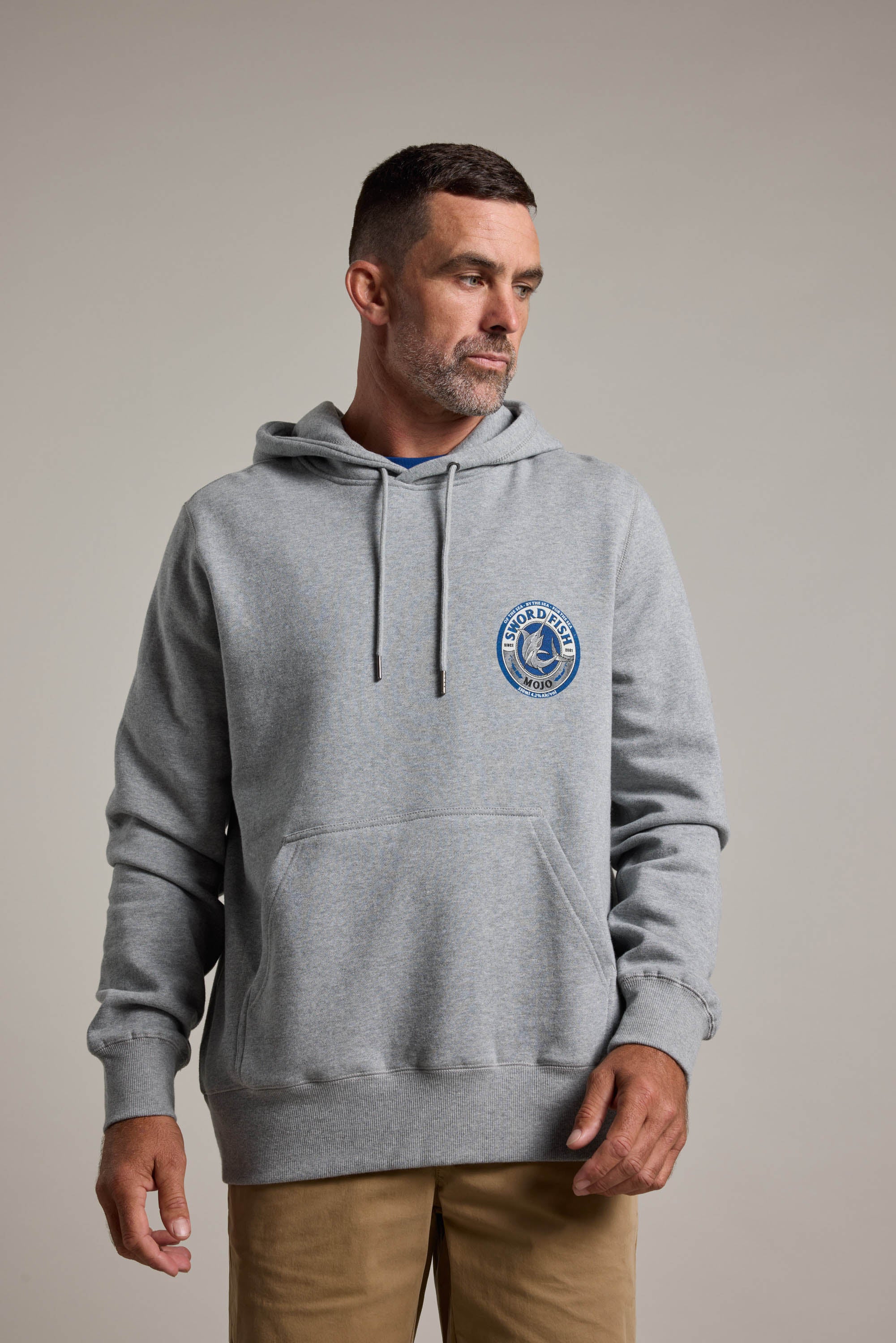 A man with short hair and a beard wears the Swordfish Mojo Logo Hoody by Swordfish Mojo in light gray, featuring a blue circular logo on the left chest, standing against a plain light background.