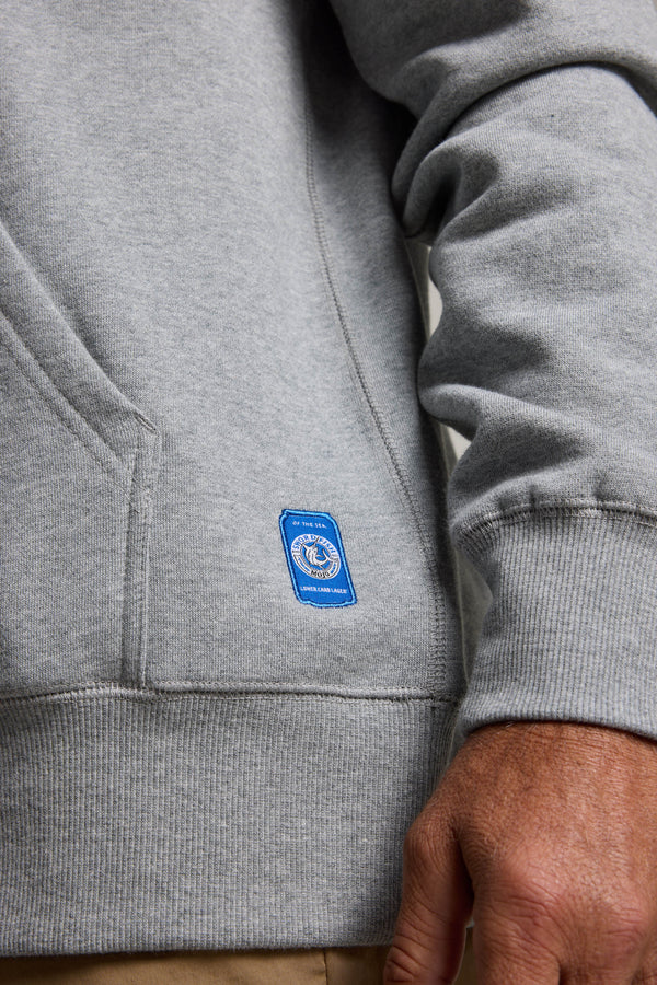 A person wearing the light gray Swordfish Mojo Logo Hoody by Swordfish Mojo, featuring a blue rectangular patch near the pocket. Their right hand is at their side, and part of a tan pant leg is visible.