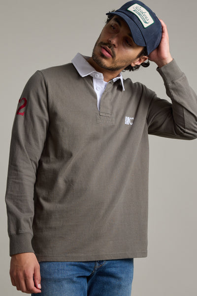 A man wearing the Barkers Stanford Long Sleeve Rugby with a white collar, blue jeans, and a dark baseball cap touches the back of his head while looking to the side against a plain background.