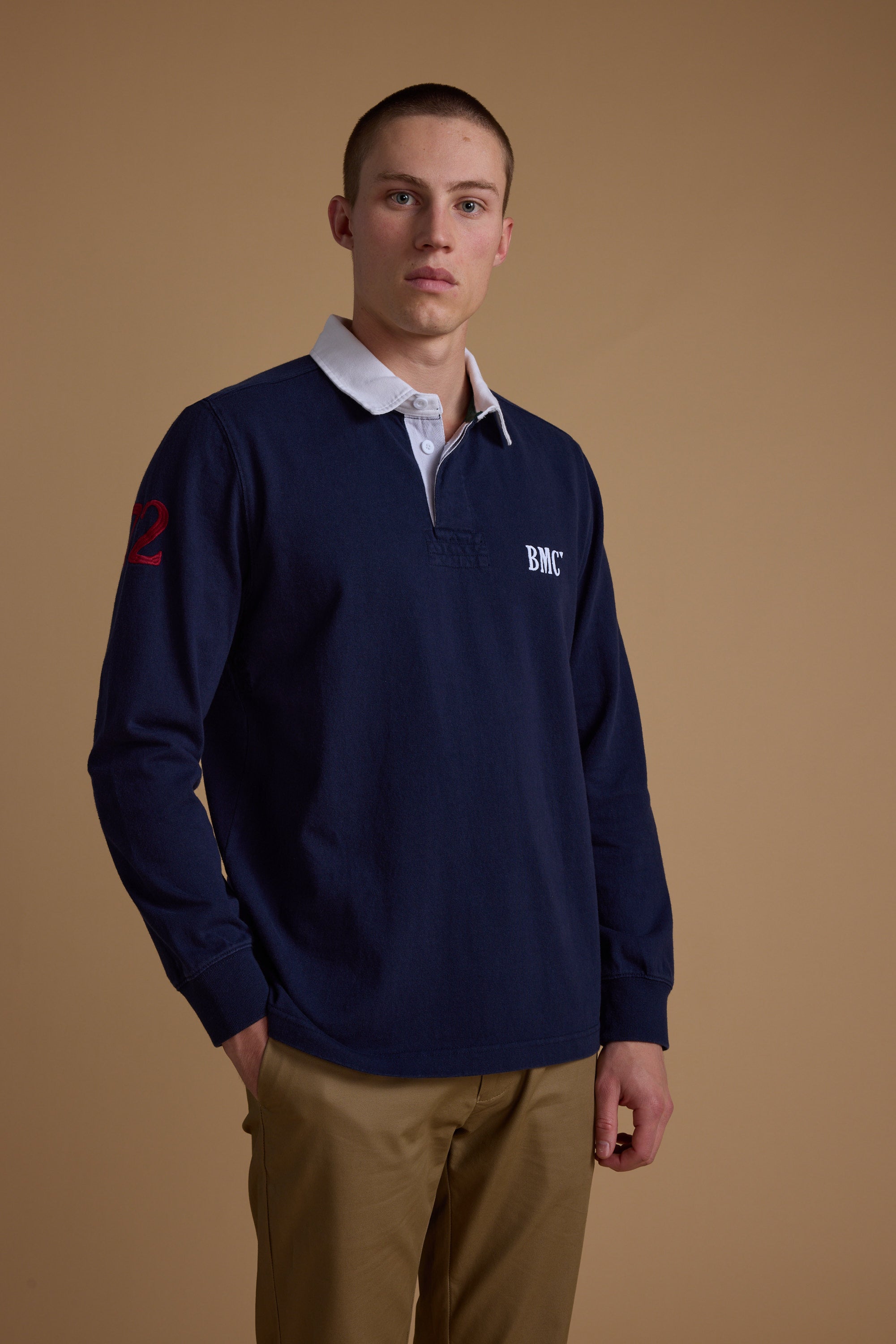 A young man with short hair wears the Barkers Stanford Long Sleeve Rugby in navy blue with a white collar and tan pants, standing before a plain brown background and looking directly at the camera.