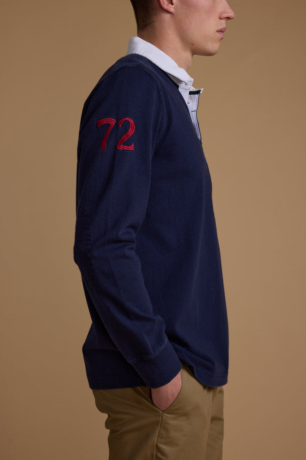 A man stands in profile against a tan background, wearing the Barkers Stanford Long Sleeve Rugby in navy with a red 72 patch on the sleeve, layered over a white collared shirt and paired with khaki pants. His hand rests in his pocket.