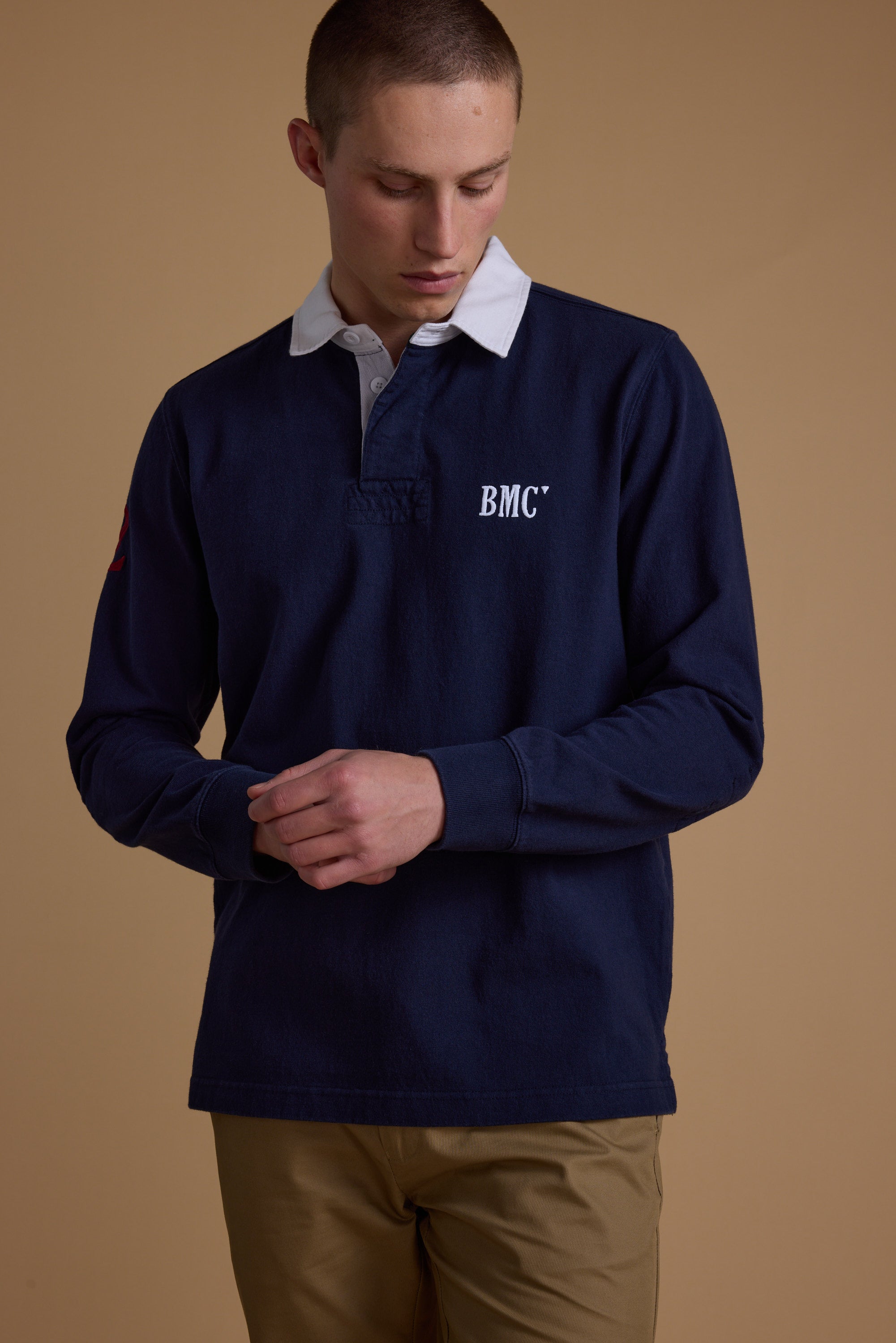 A person in a Barkers Stanford Long Sleeve Rugby—navy organic cotton with a white collar and BMC text—stands against a tan background, looking down with hands lightly clasped.