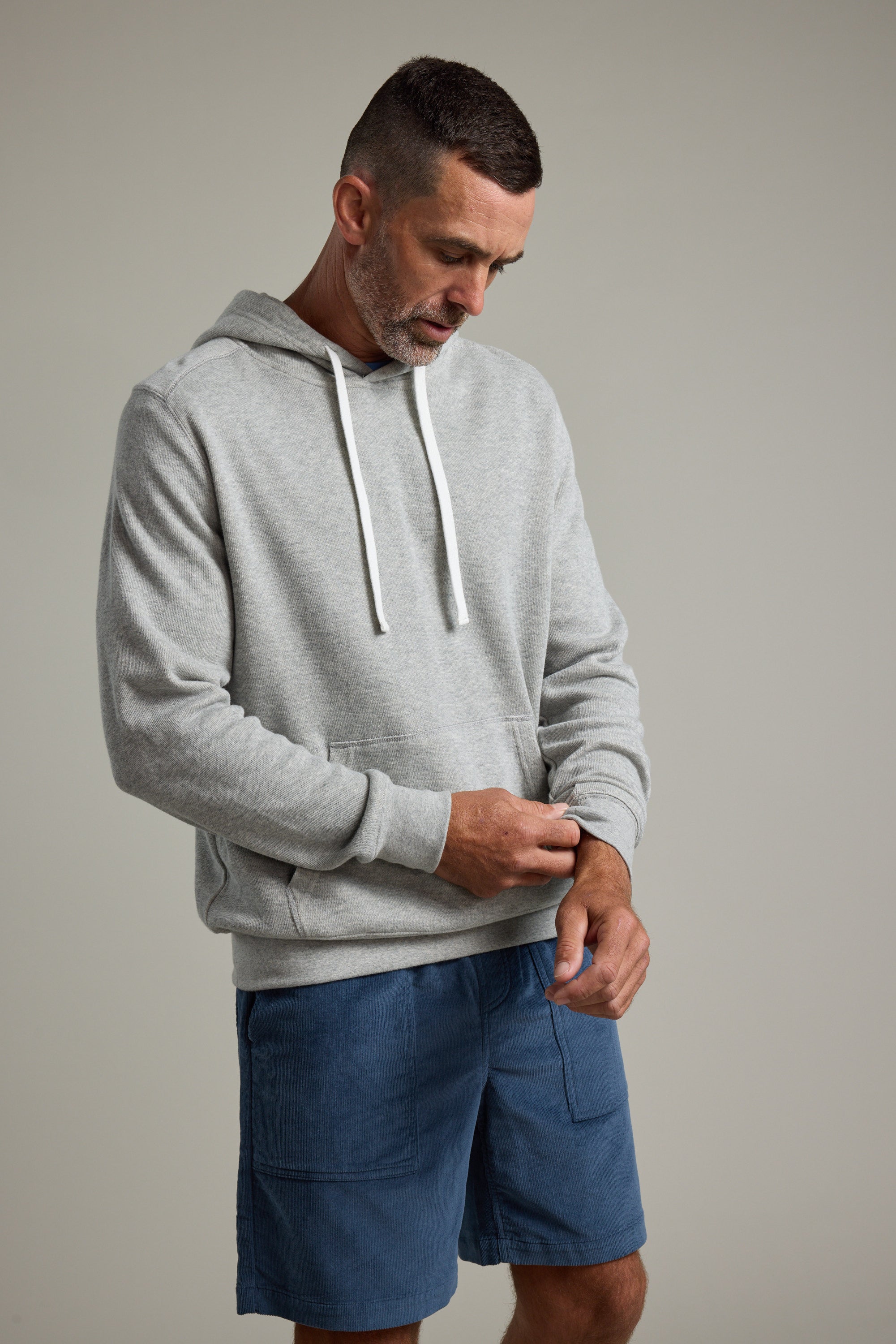 A man wearing the Barkers Inlet Hoody in light gray and blue shorts looks down, adjusting his sleeve against a plain, light background.