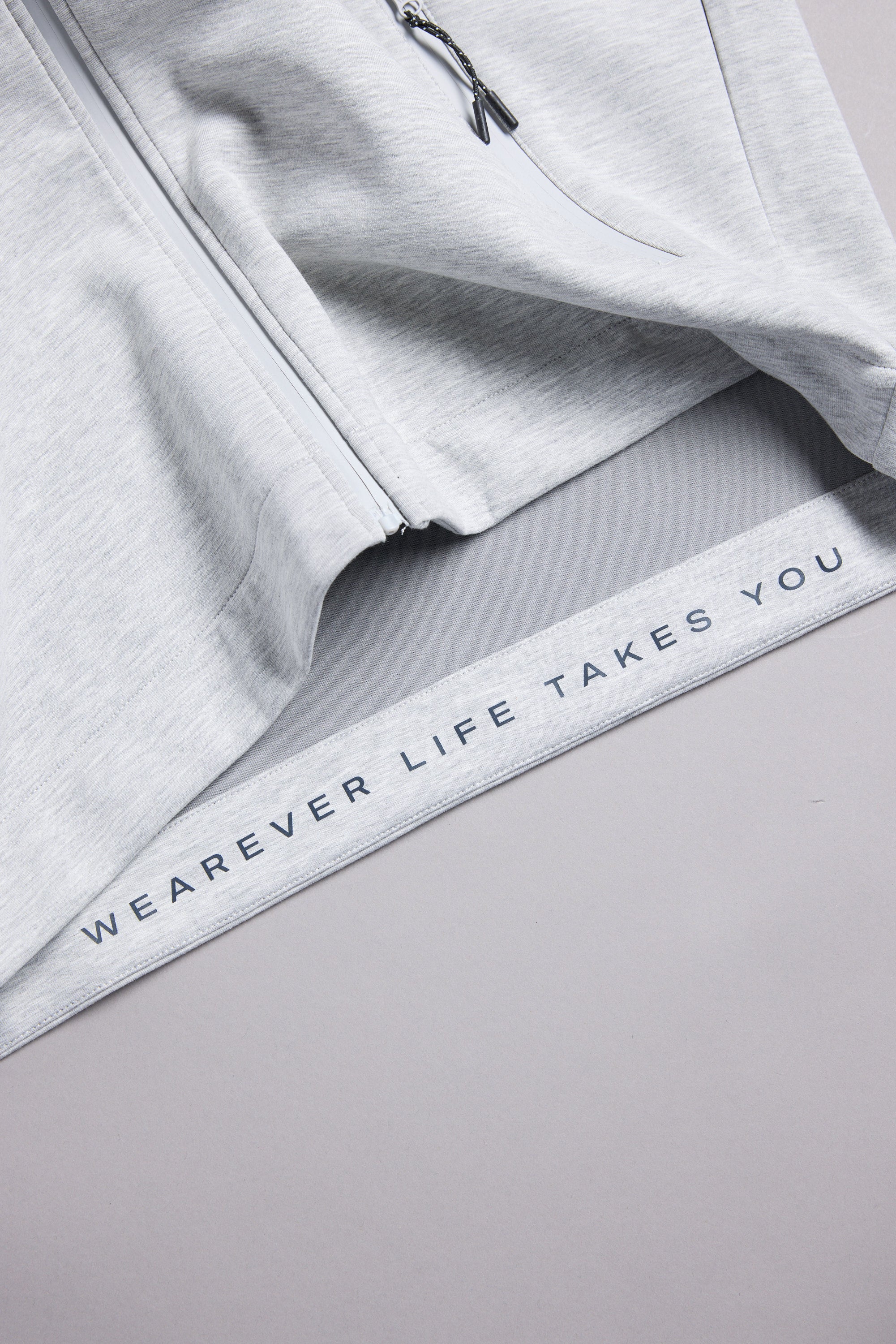 A close-up of the WearEver Altitude Hoody, a light gray tech fleece zip-up by WearEver, featuring "WEAREVER LIFE TAKES YOU" printed along the inner hem.
