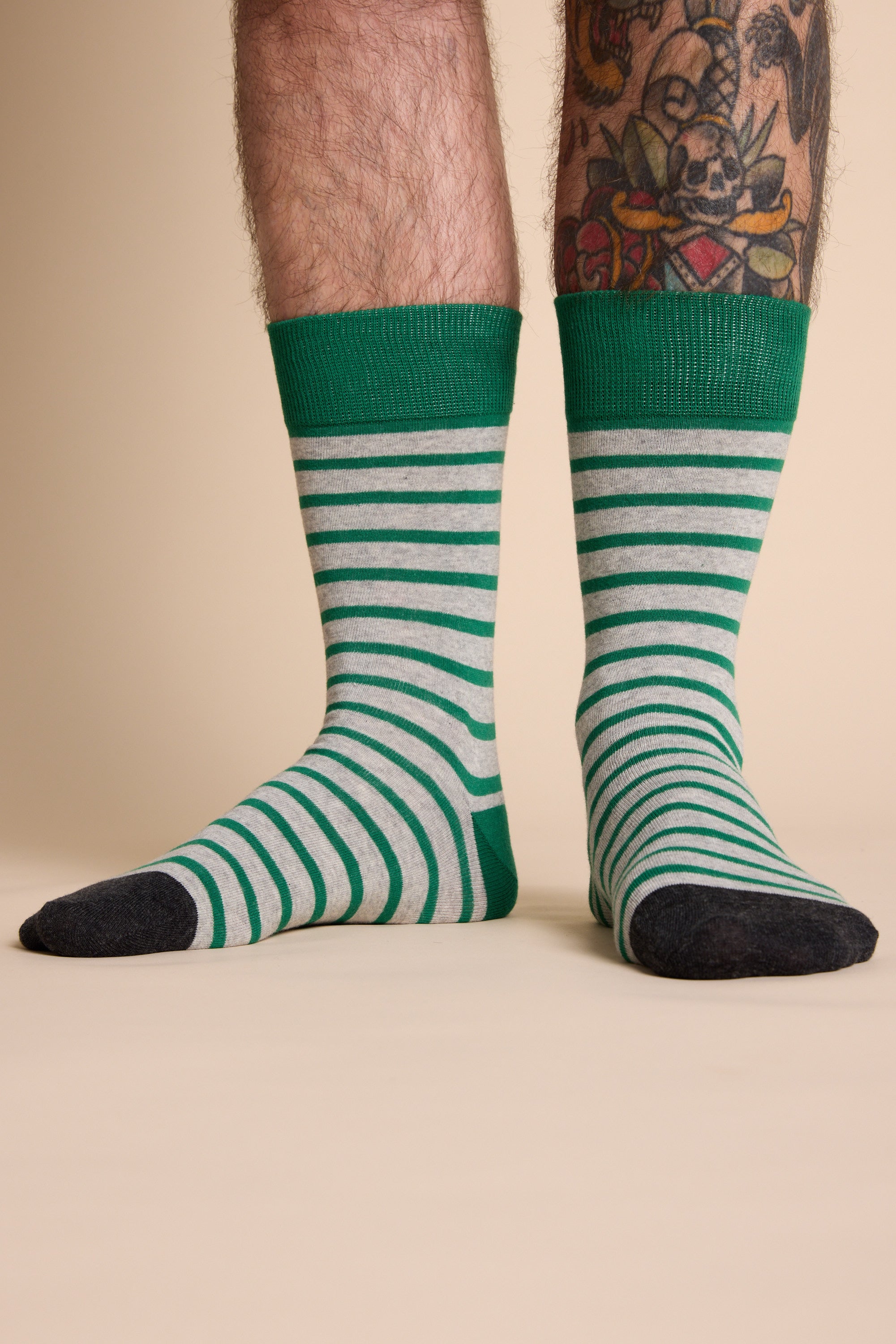 A person with tattooed legs wears Barkers Mojave Stripe Sock—green and white cotton socks with dark toes and heels—while standing on a beige surface against a neutral background.